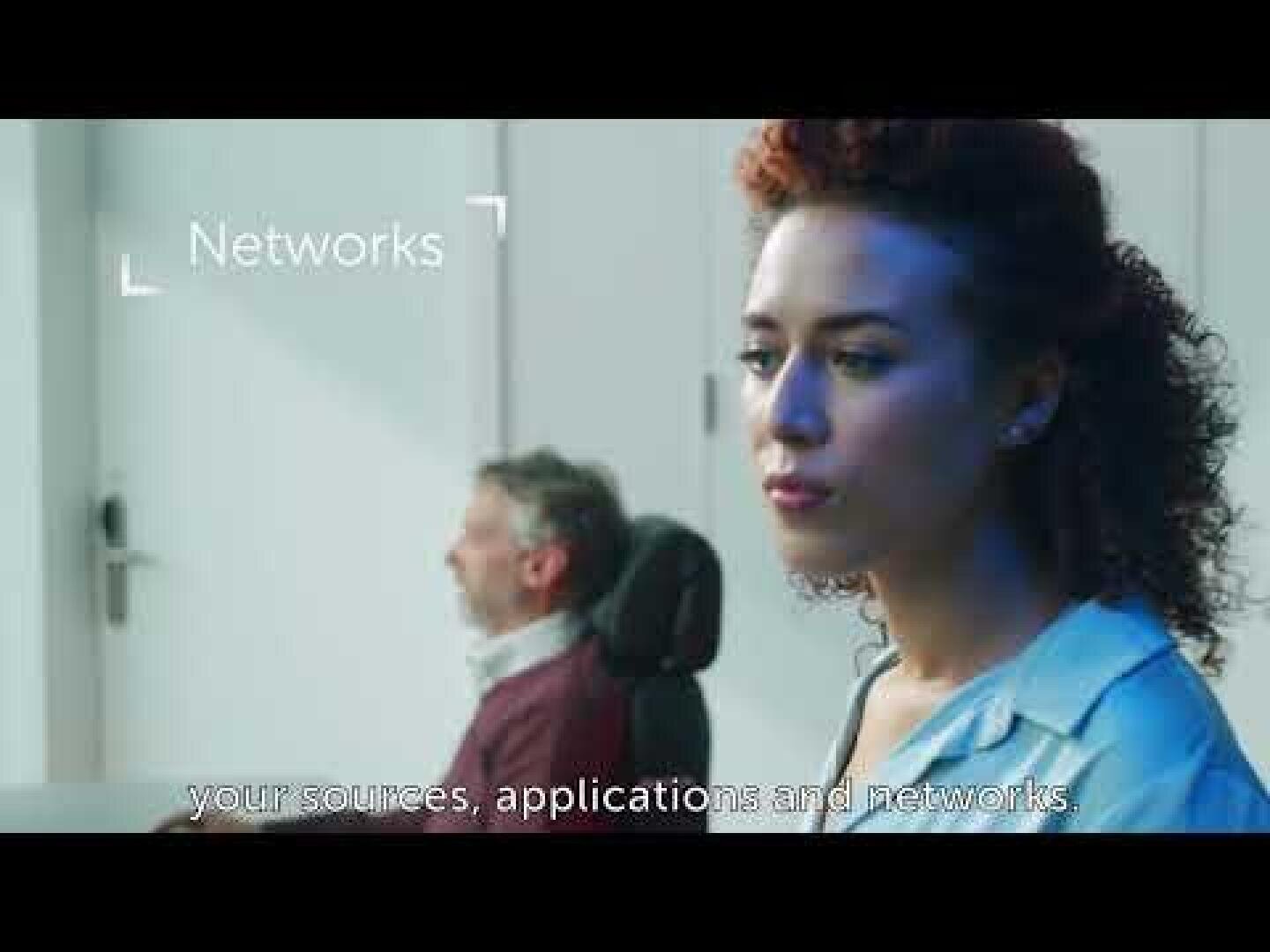 Eine Frau mit lockigem Haar blickt konzentriert in den Vordergrund, während ein Mann im Hintergrund sitzt. Auf dem Bildschirm erscheint das Wort "Networks" (Netzwerke) zusammen mit Untertiteln, in denen Ihre Quellen, Anwendungen und Netzwerke zu lesen sind.