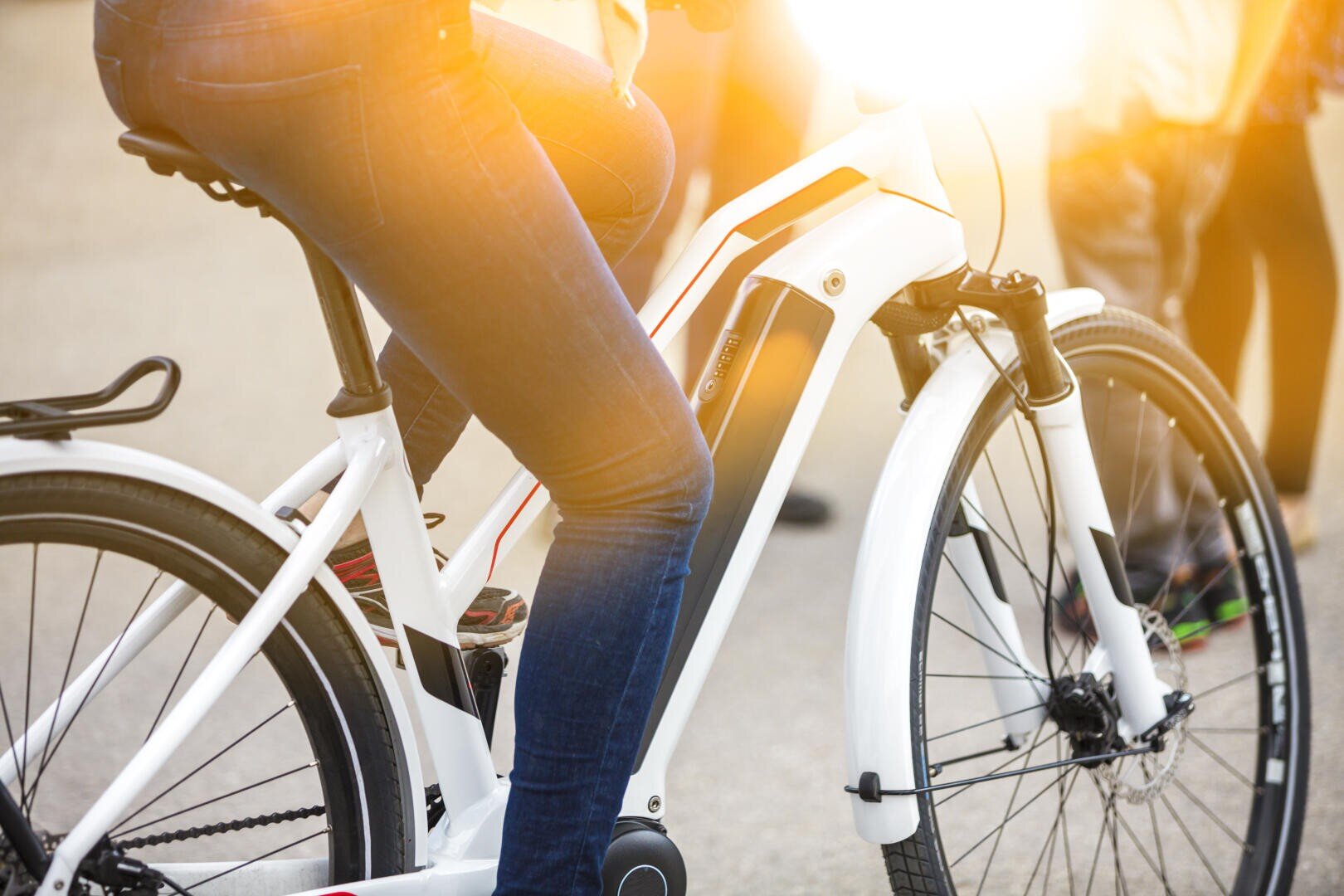 Eine Person in Jeans sitzt draußen auf einem weißen E-Bike, im Hintergrund sind andere Menschen verschwommen zu sehen, und Sonnenlicht fällt auf die Szene.