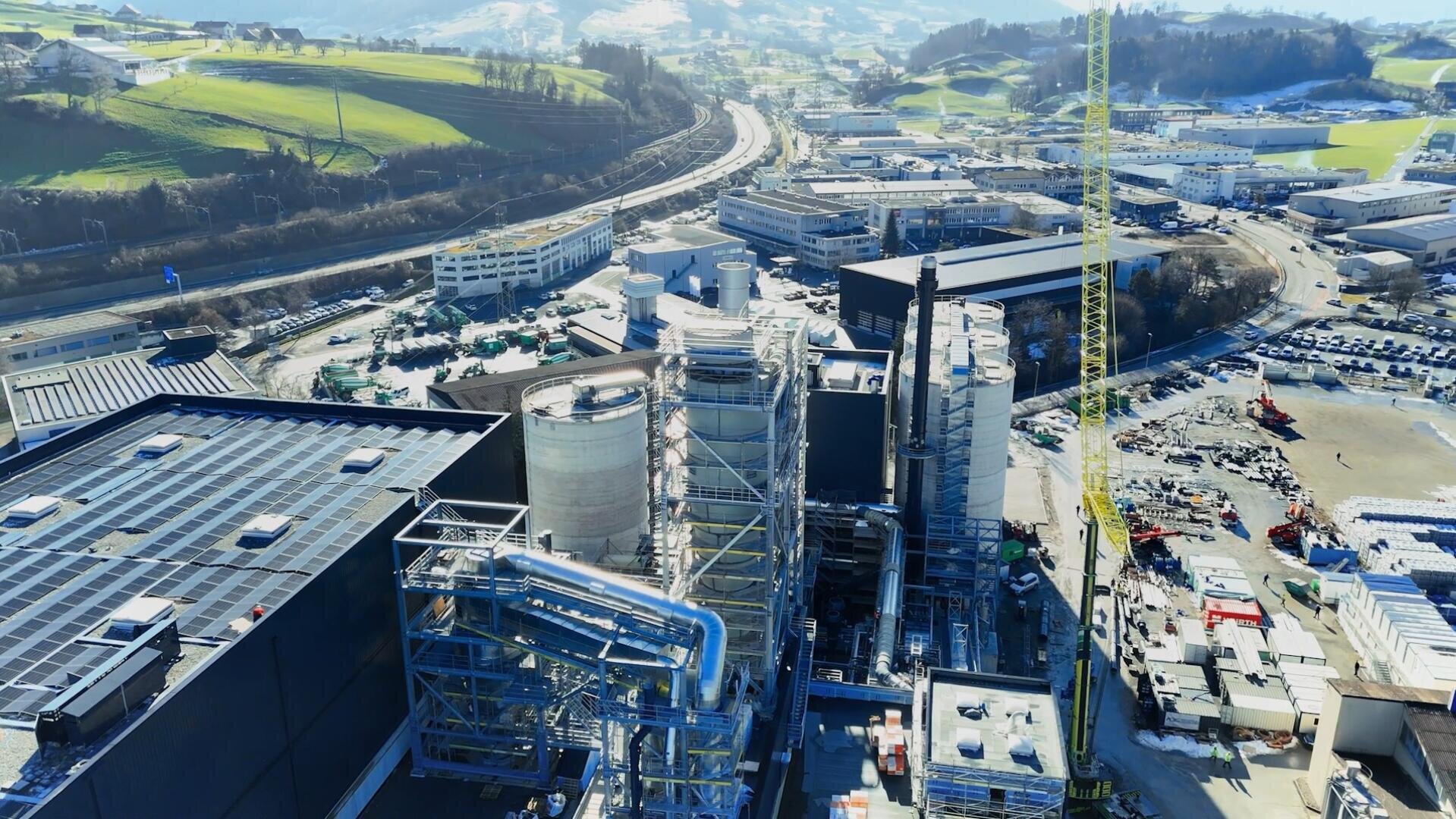 Luftaufnahme einer Industrieanlage mit großen Tanks, Rohren und Strukturen, umgeben von Straßen, Gebäuden, geparkten Fahrzeugen und grünen Hügeln unter einem klaren Himmel.