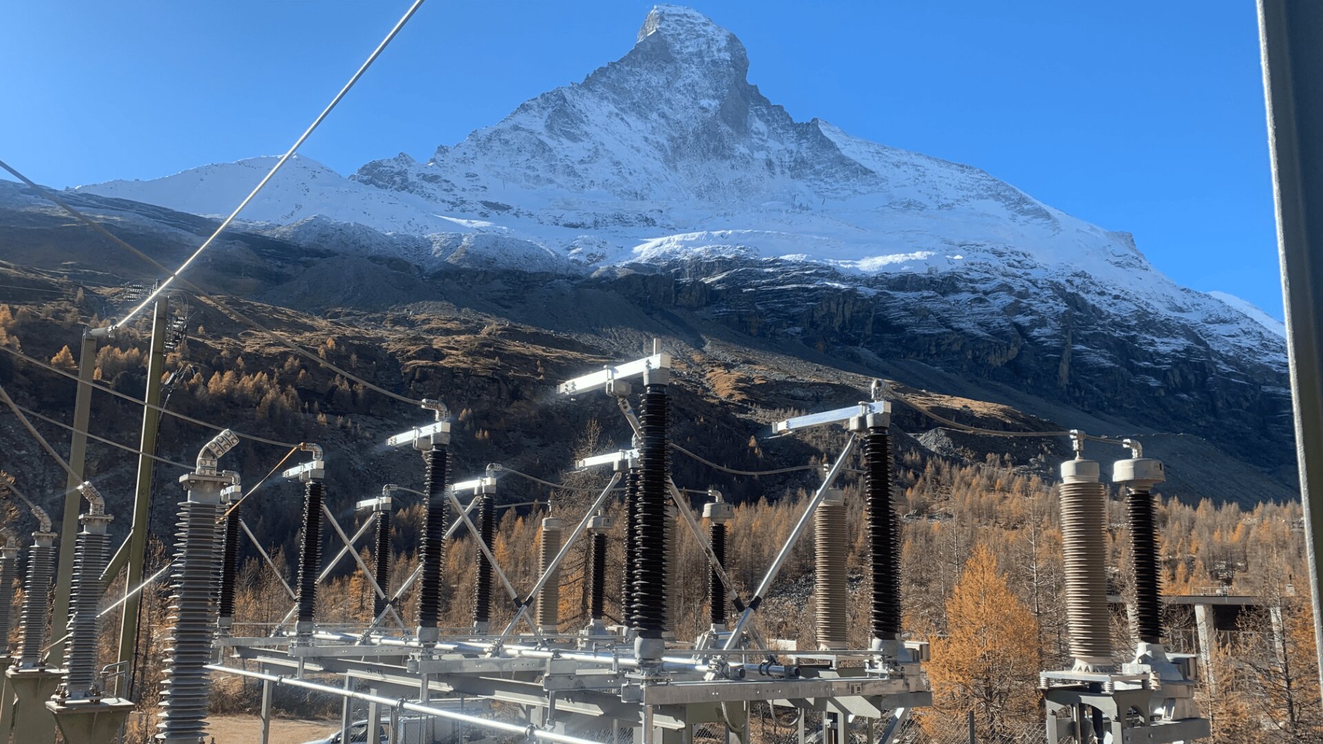 Komponenten eines Umspannwerks mit Metall- und Keramikisolatoren im Vordergrund, vor dem Hintergrund von Herbstbäumen und einem schneebedeckten Berggipfel unter blauem Himmel.