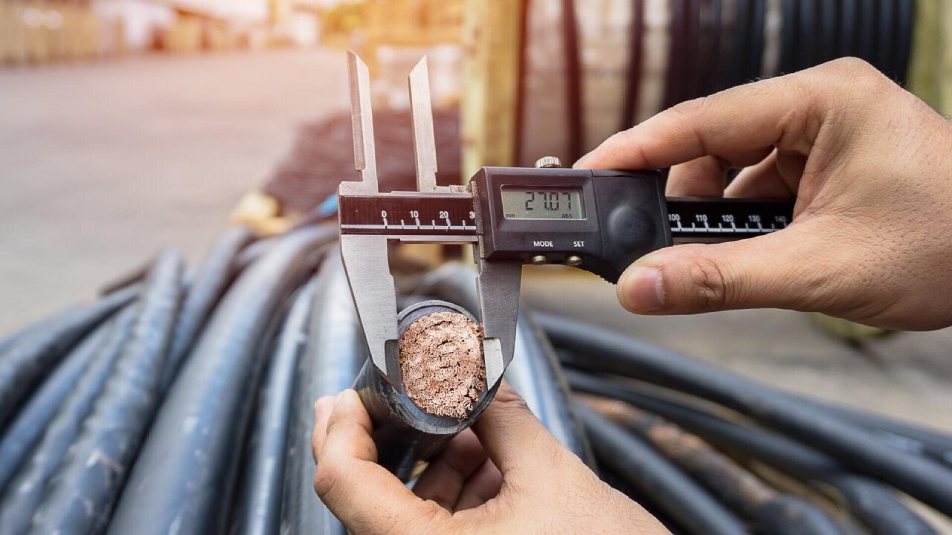 A person uses a digital caliper to measure the diameter of a thick, cut electrical cable, revealing exposed copper wires inside. Coiled cables are visible in the background.