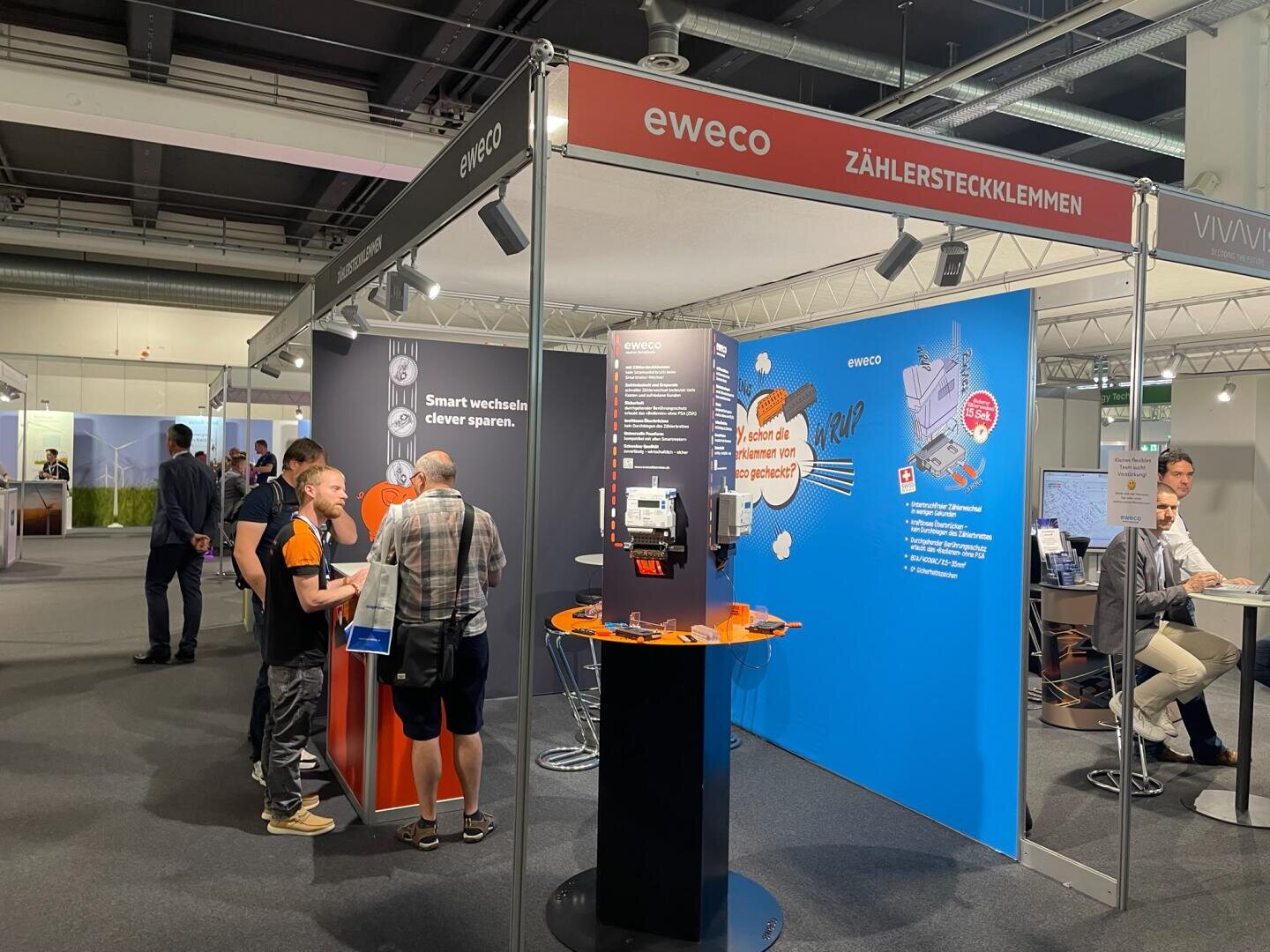 Der Messestand von eWeco umfasst Informationsdisplays, einen zentralen orange-blauen Stand, Werbematerialien sowie Besucher, die sich in einer hell beleuchteten Messehalle mit den Mitarbeitern austauschen.