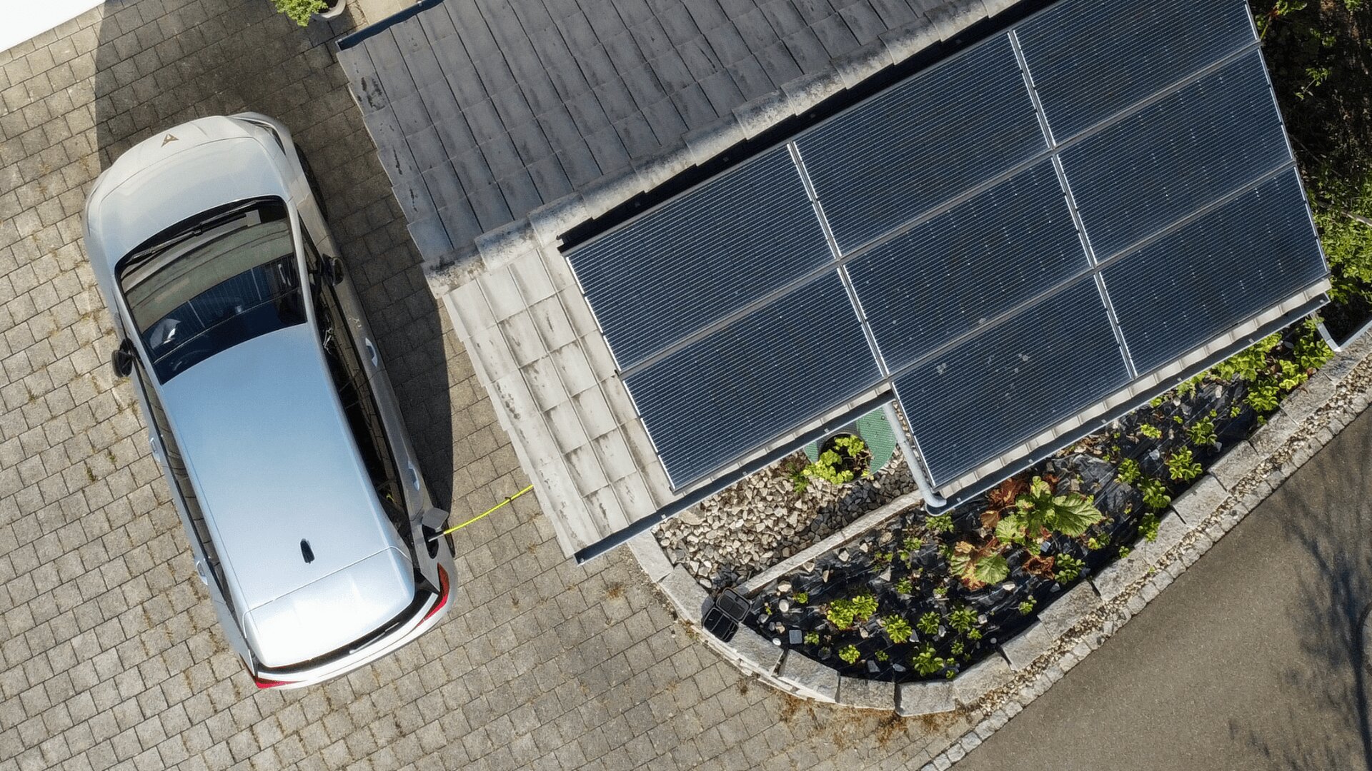 Ein silbernes Elektroauto ist auf einer Auffahrt geparkt, angeschlossen an eine Ladestation neben einem Gebäude mit Sonnenkollektoren auf dem Dach und einem begrünten Garten daneben, von oben gesehen.