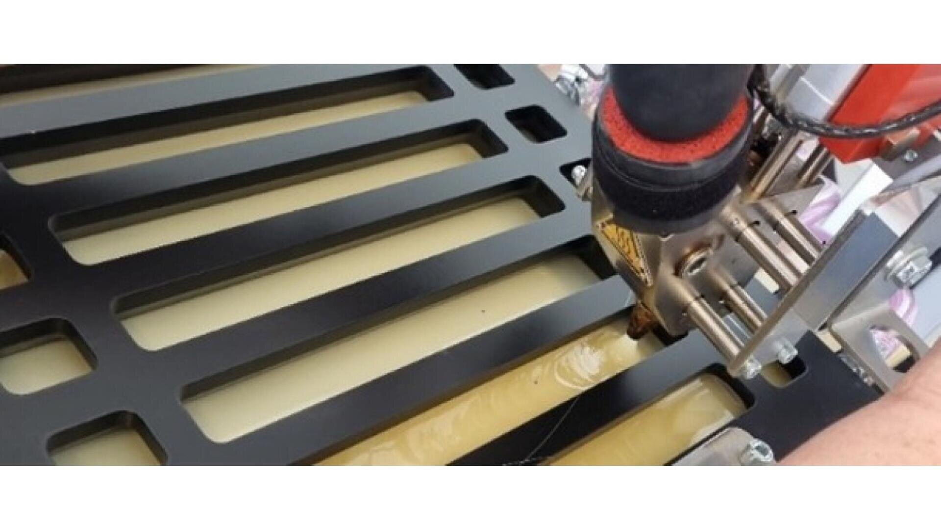 A close-up of a CNC machine or 3D printer extruding material onto a slotted metal surface, with a hand visible on the right side guiding the process.