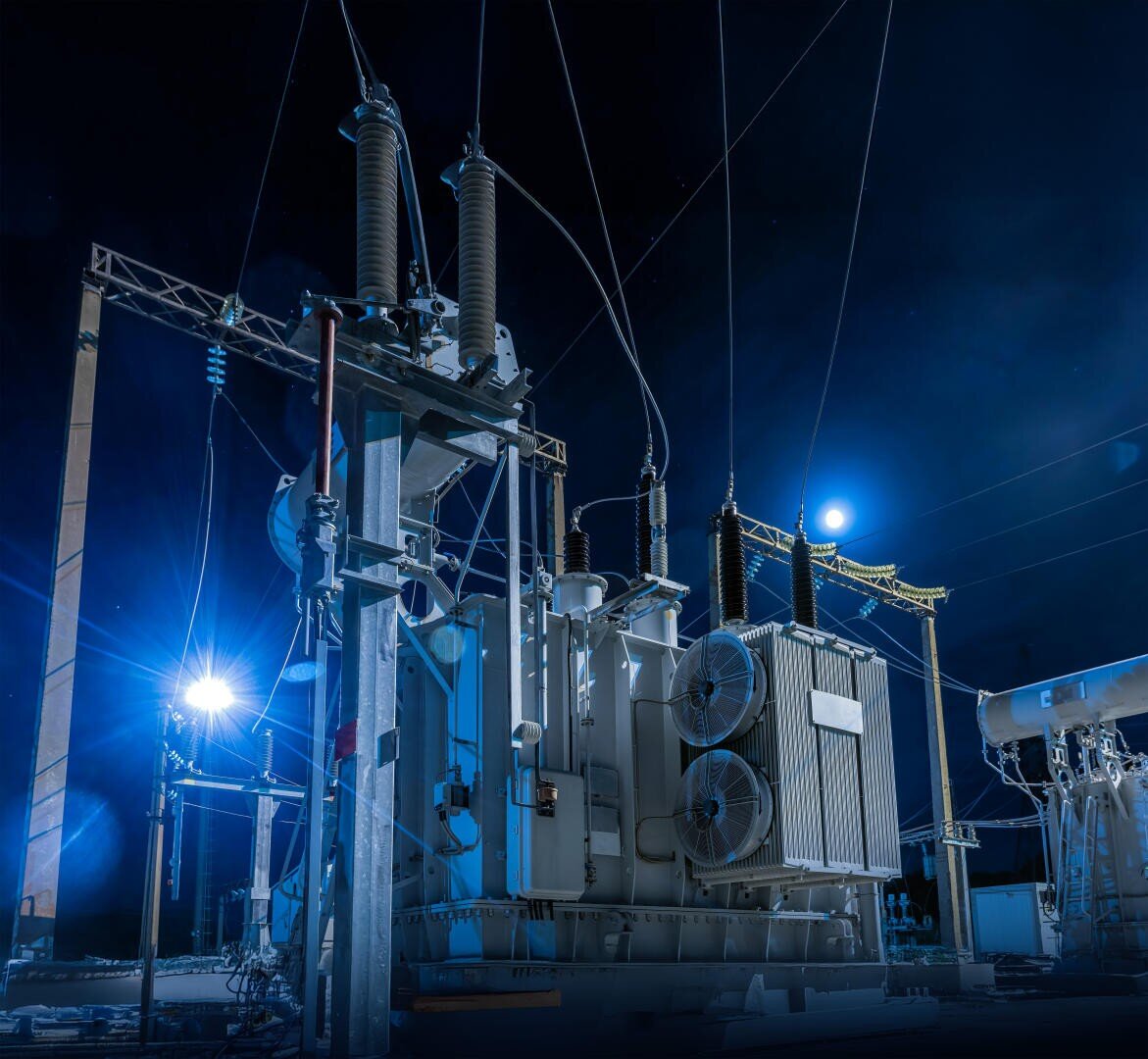 Ein nächtliches Hochspannungsumspannwerk, blau beleuchtet, mit großen Transformatoren, Isolatoren und Stromleitungen vor einem dunklen Himmel, in dem der Mond im Hintergrund zu sehen ist.