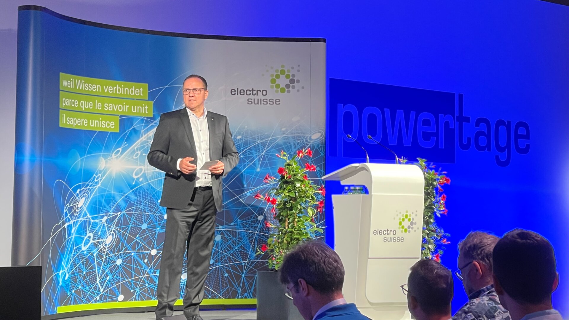 A man in a suit stands on stage delivering a speech at an Electro Suisse event. He is beside a podium with flowers. The background features digital graphics and the word powertage in large blue letters.