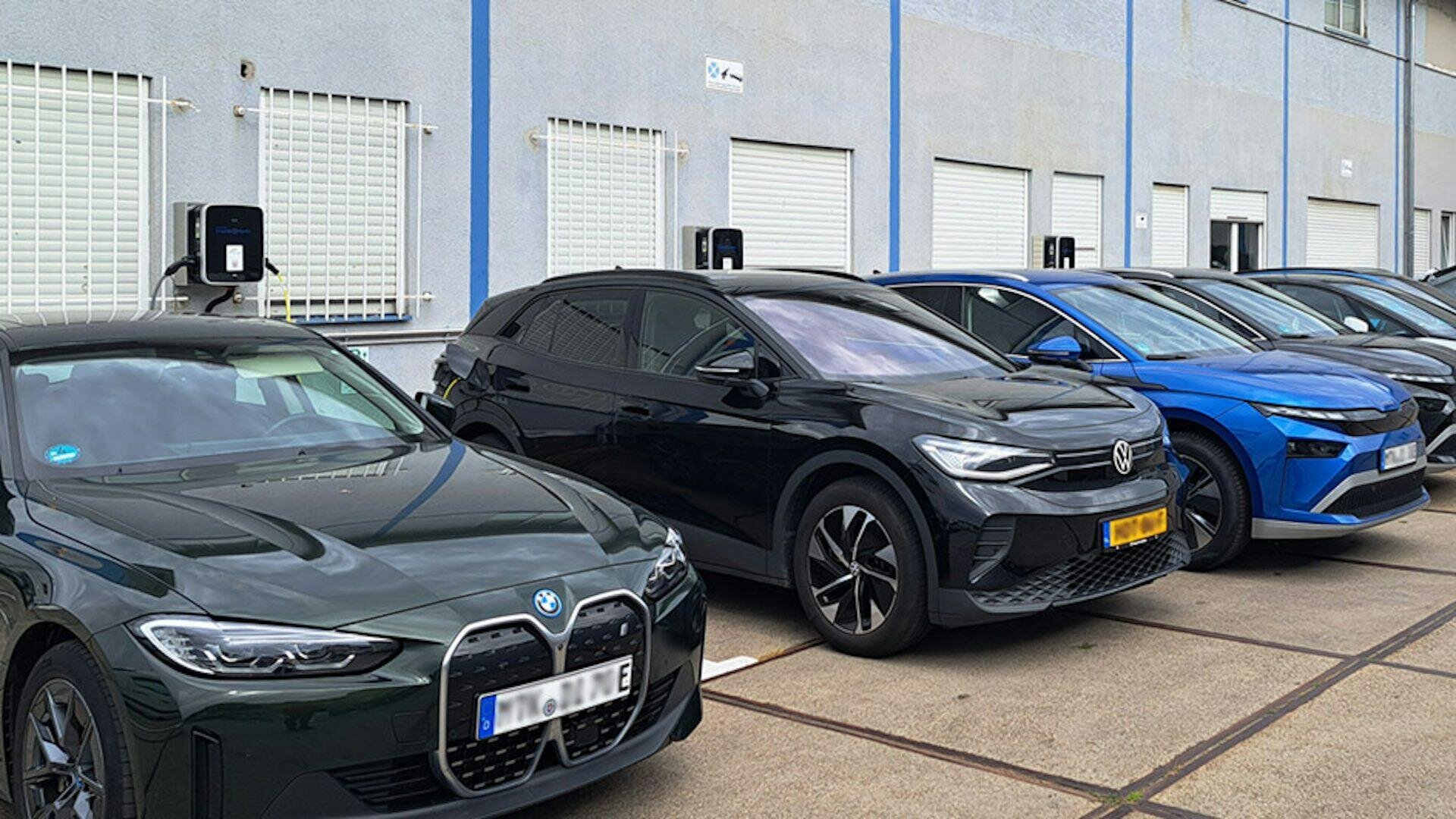 Mehrere Elektroautos, darunter ein BMW und ein Volkswagen, sind nebeneinander geparkt und laden an Wandladestationen vor einem Gebäude mit weißen Fensterläden.