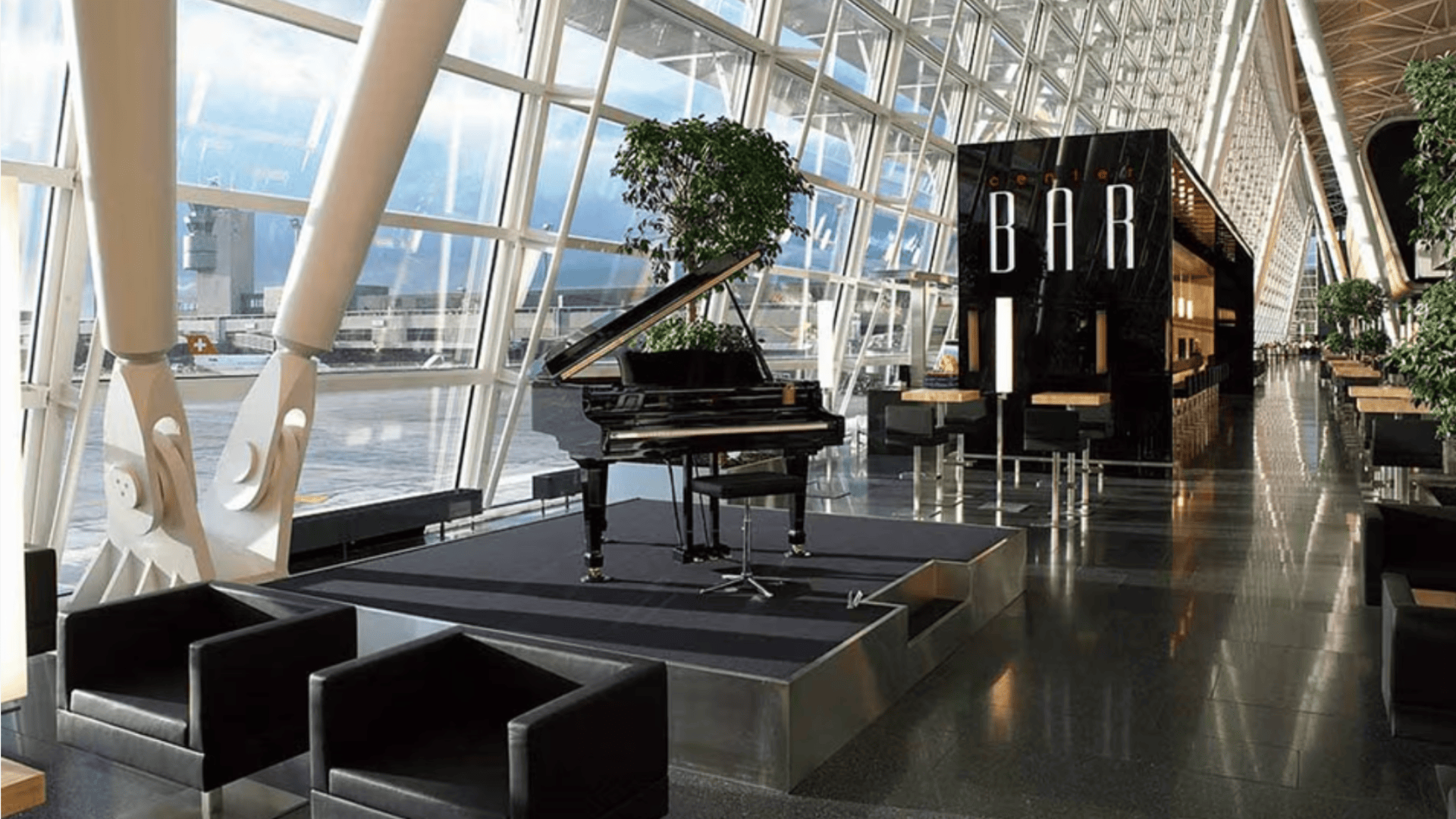 Ein Flügel steht auf einem erhöhten Podest in einem modernen, geräumigen Flughafenterminal mit großen Fenstern, schwarzen Stühlen, Zimmerpflanzen und einer Bar im Hintergrund.