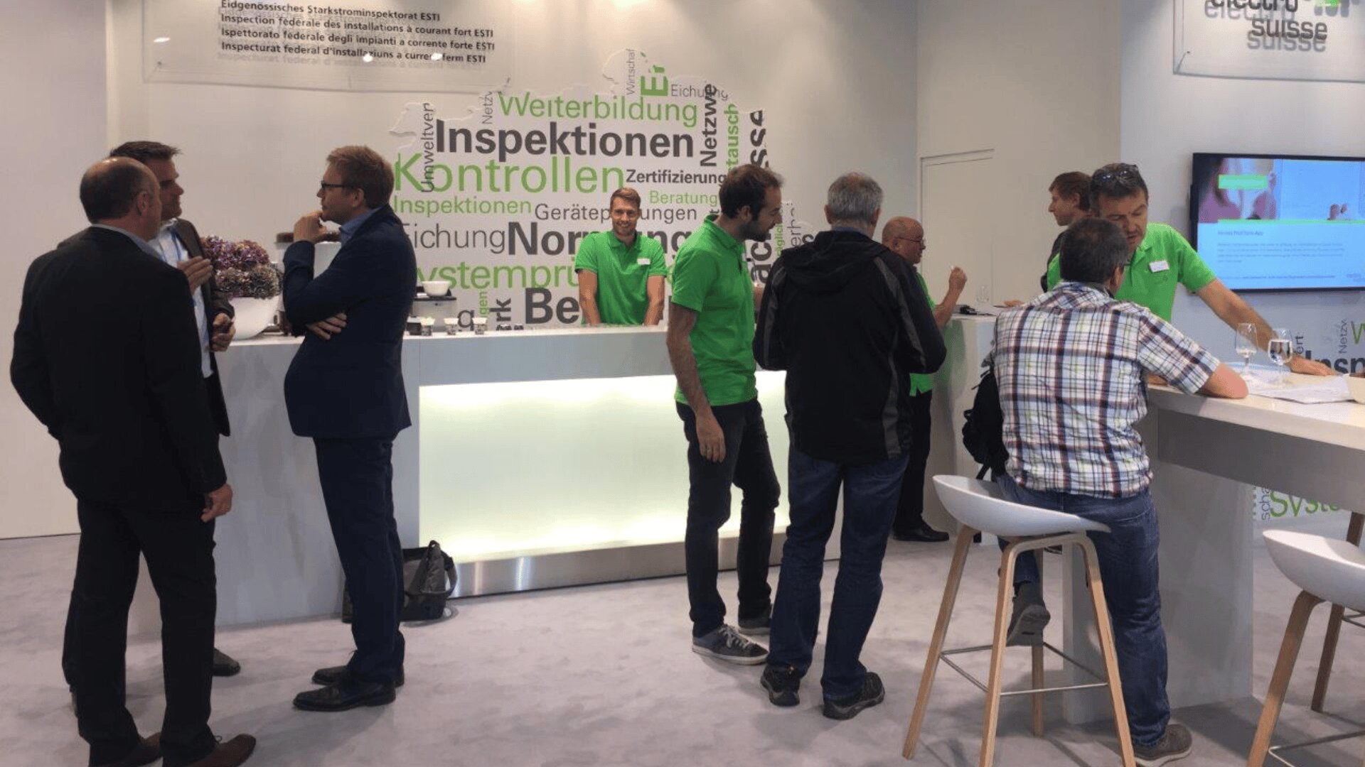 Eine Gruppe von Männern, einige in grünen Hemden, stehen und unterhalten sich an einem modernen Messestand mit weißem Tresen. An der Rückwand sind deutsche Worte zu lesen, und auf der rechten Seite ist ein Bildschirm zu sehen.