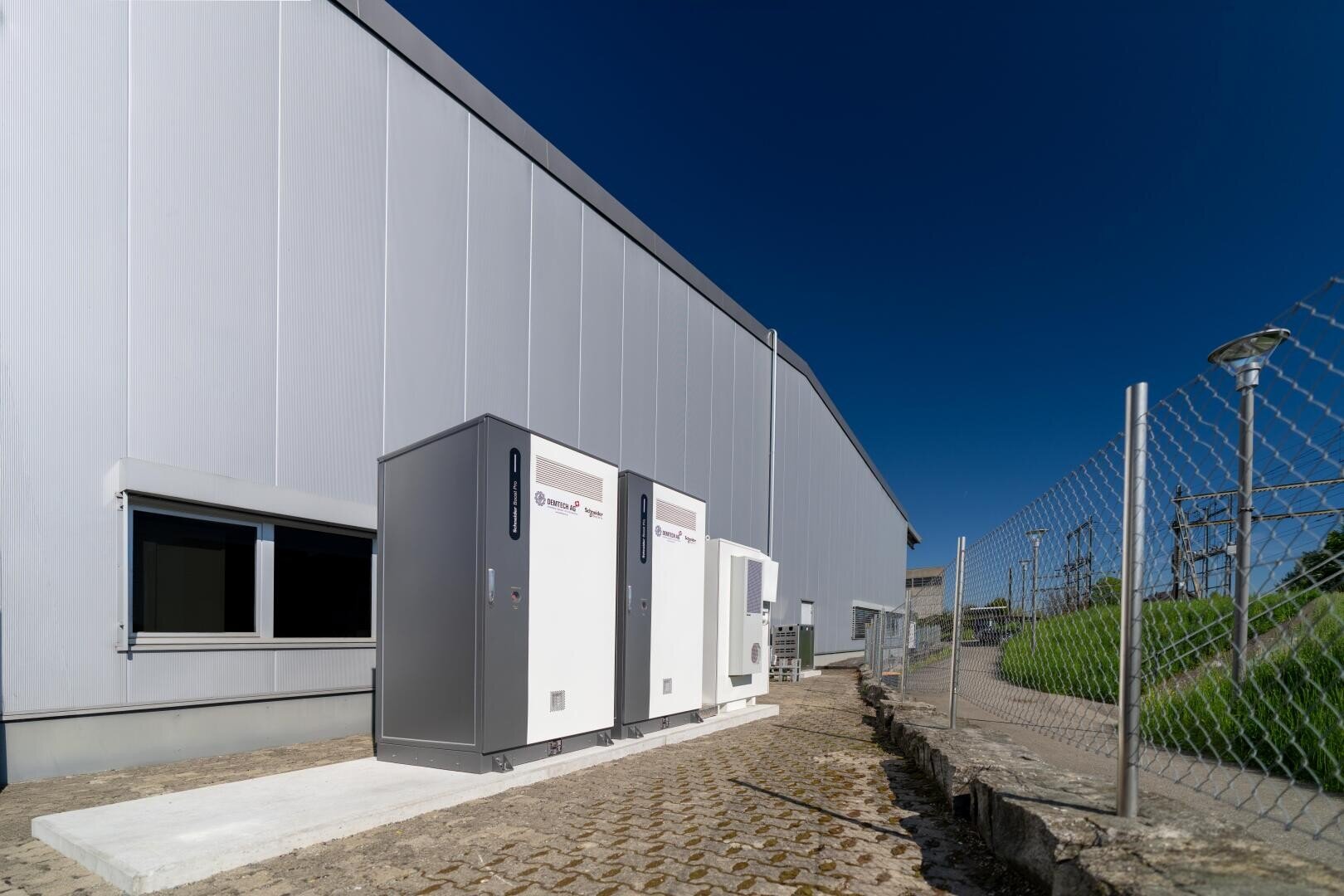 Drei industrielle Schaltschränke sind im Freien auf einer Betonfläche neben einem großen grauen Gebäude installiert, mit einem Maschendrahtzaun und einem klaren blauen Himmel im Hintergrund.