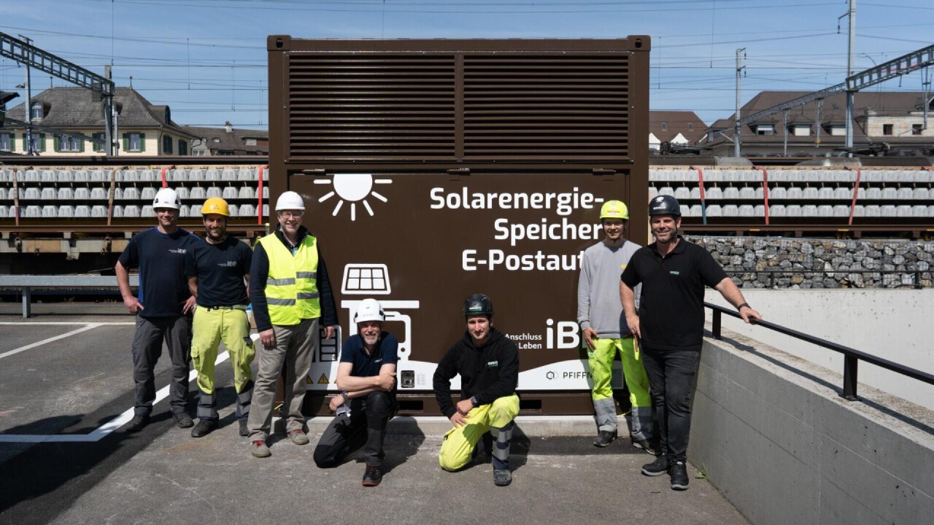 Seven workers in safety gear stand and kneel in front of a large brown container labeled Solarenergie-Speicher E-Postauto, with rail tracks and buildings in the background on a sunny day.