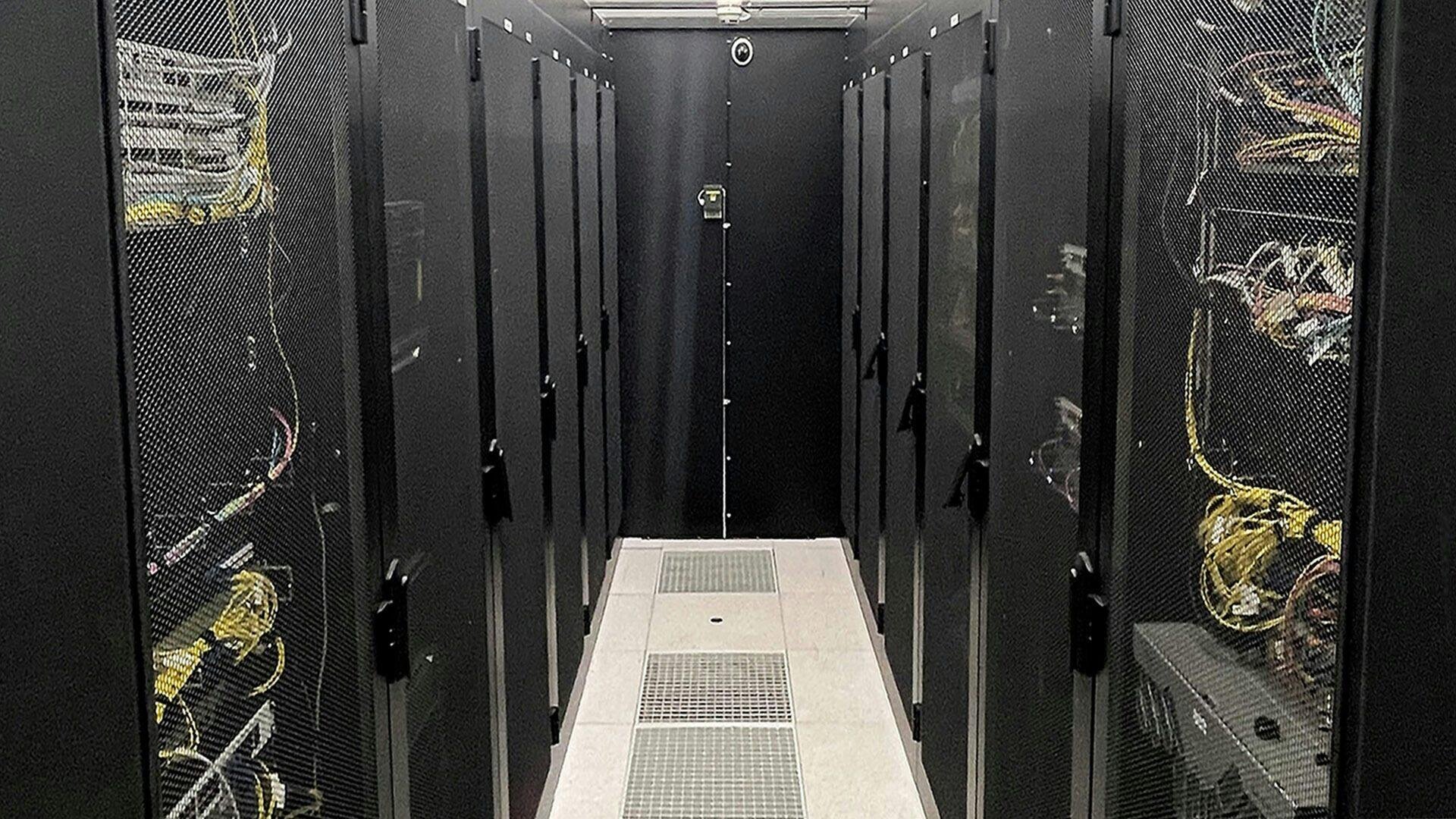 A server room with rows of black, mesh-fronted server racks on both sides, filled with cables and equipment. The aisle is lit and has vents on the floor, with a security camera visible at the end of the aisle.