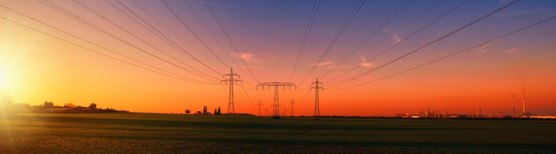 Sonnenuntergangsansicht von Stromleitungen, die sich über eine grüne Wiese erstrecken, mit Strommasten, die in Richtung eines Industriegebiets am Horizont unter einem bunten Himmel führen.