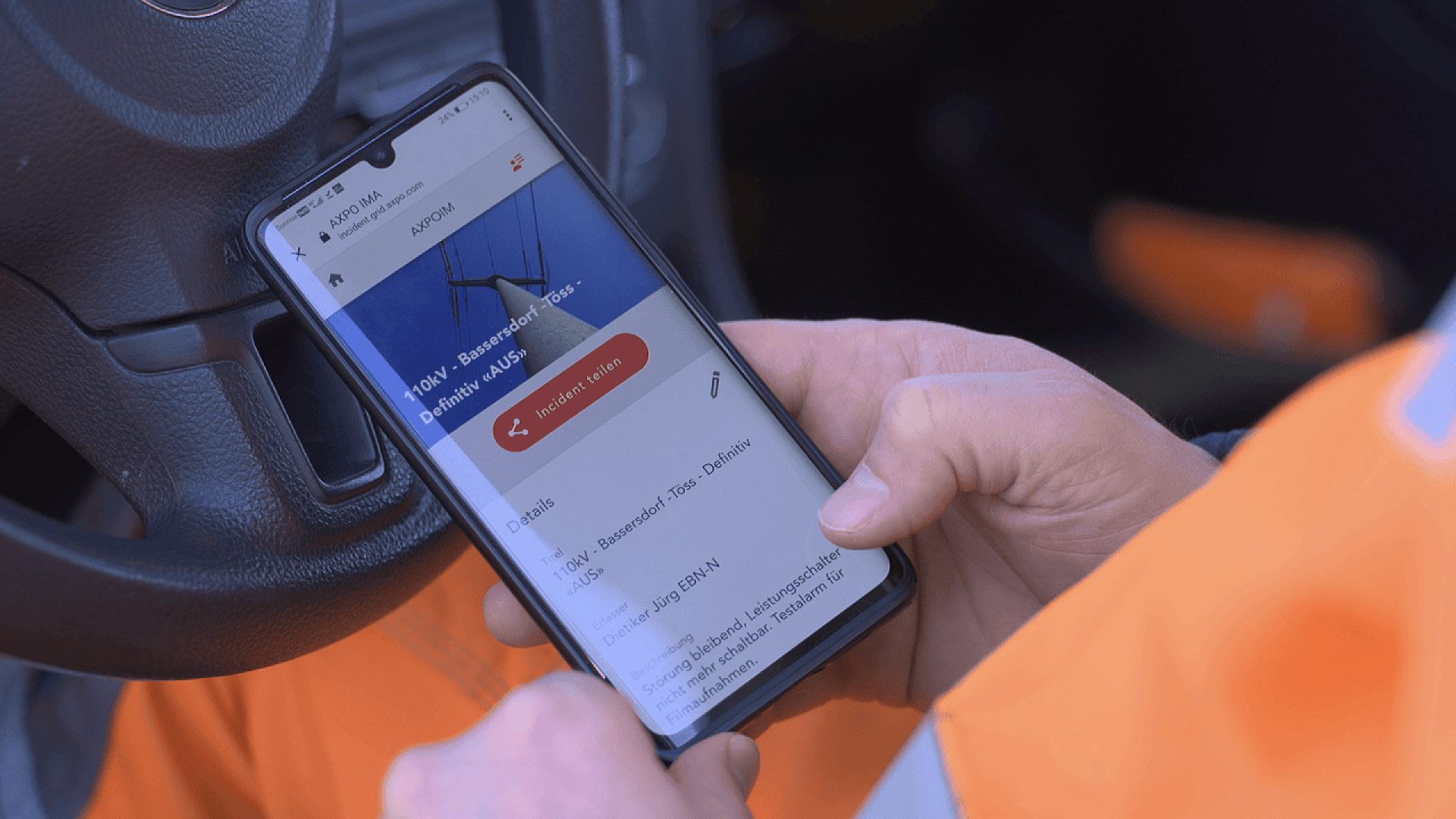 A person wearing an orange jacket uses a smartphone inside a vehicle, viewing an app screen with instructions and information displayed.