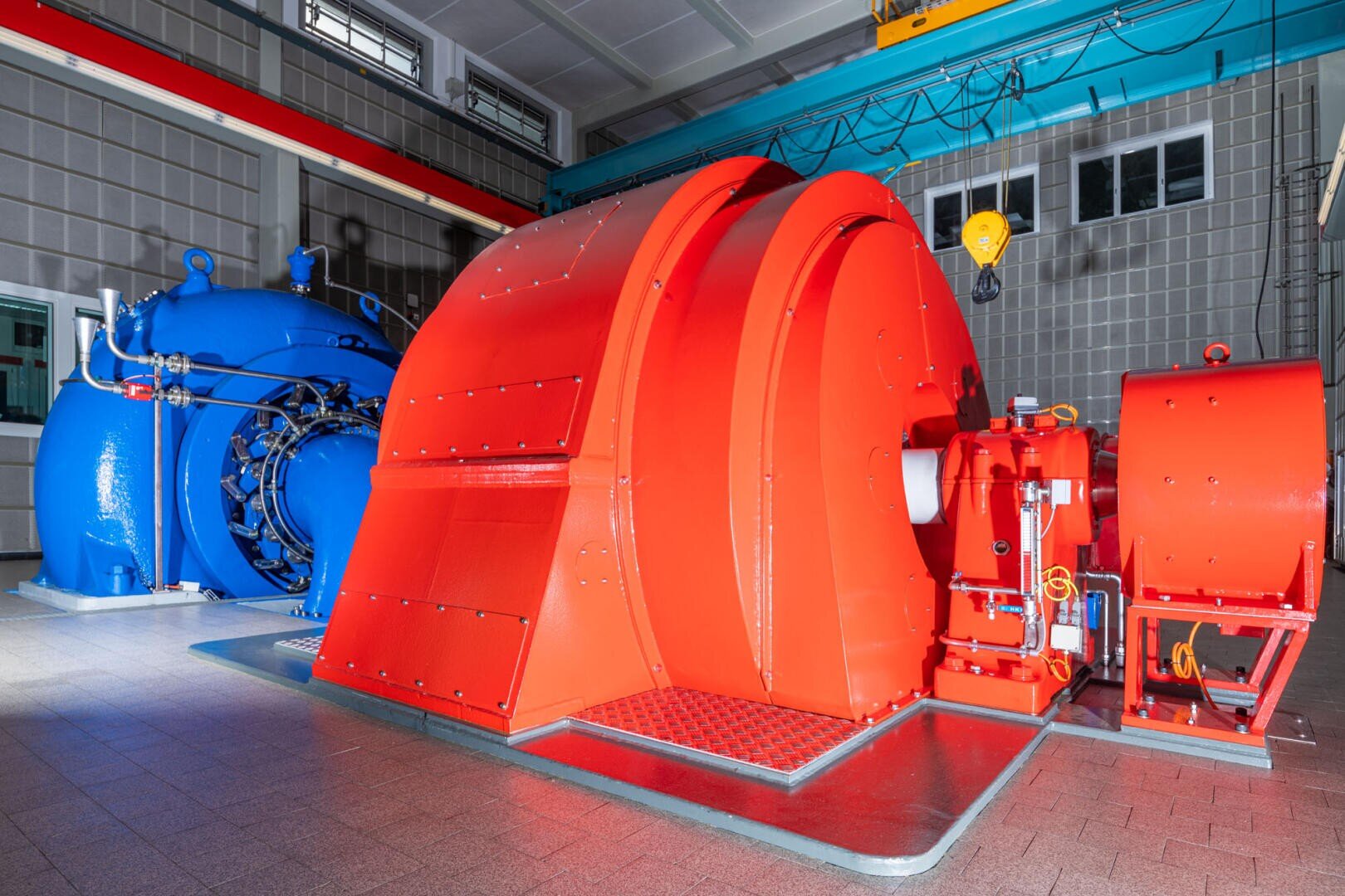 Große Industriemaschinen im Inneren einer Anlage, mit einem auffälligen roten Generator oder einer Turbine im Vordergrund und einem blauen Generator oder einer Turbine dahinter, mit gekachelten Wänden und sichtbaren Rohren und Kabeln.
