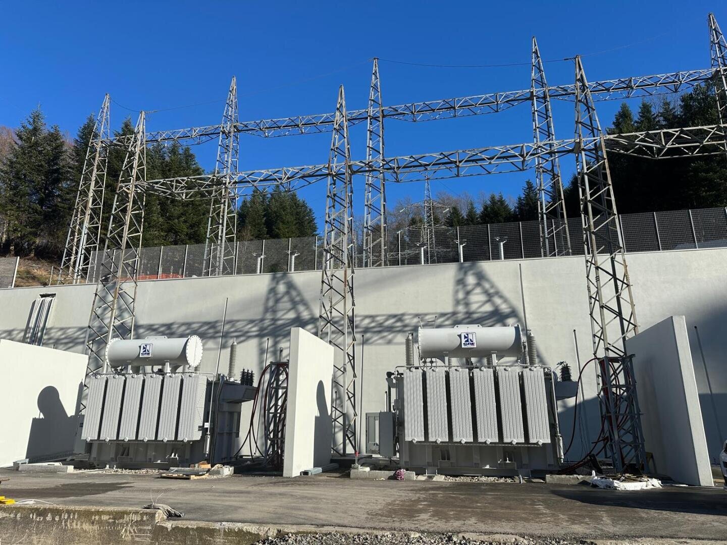 Two large electrical transformers installed in an outdoor substation, featuring metal lattice structures and overhead power lines with trees and a clear blue sky in the background.
