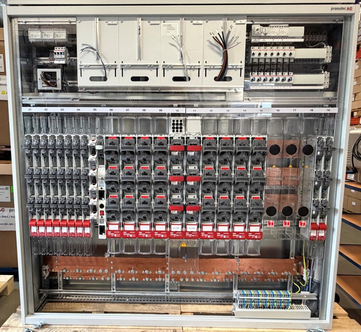 Industrielle elektrische Schalttafel mit mehreren Leistungsschaltern, Relais, Sicherungen, Verdrahtungsklemmen und beschrifteten Modulen, die übersichtlich in einem Metallgehäuse untergebracht sind. Einige Drähte sind noch nicht angeschlossen.