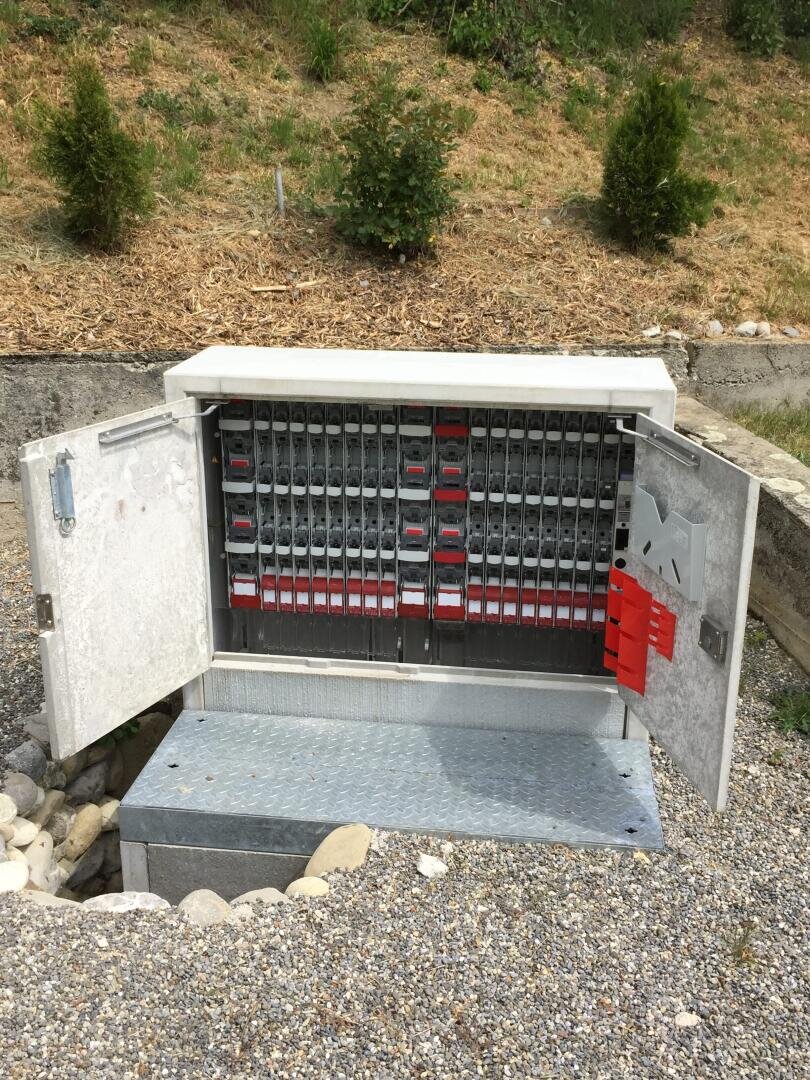 Ein offener Schaltschrank im Freien mit mehreren Reihen von Schaltern und Bauteilen auf einer Kiesfläche mit kleinen Büschen und grasbewachsenem Boden im Hintergrund.