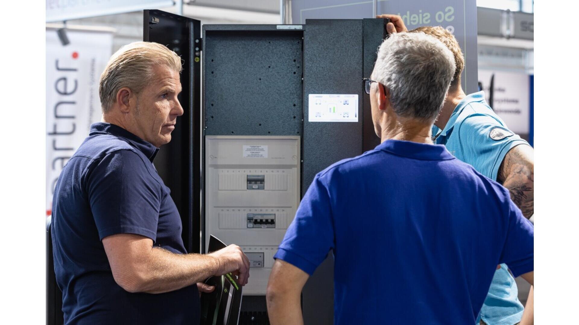 Three men stand in front of an open electrical panel, discussing its contents. Two face the panel while one gestures towards it, suggesting a technical conversation or equipment demonstration at an event or workplace.