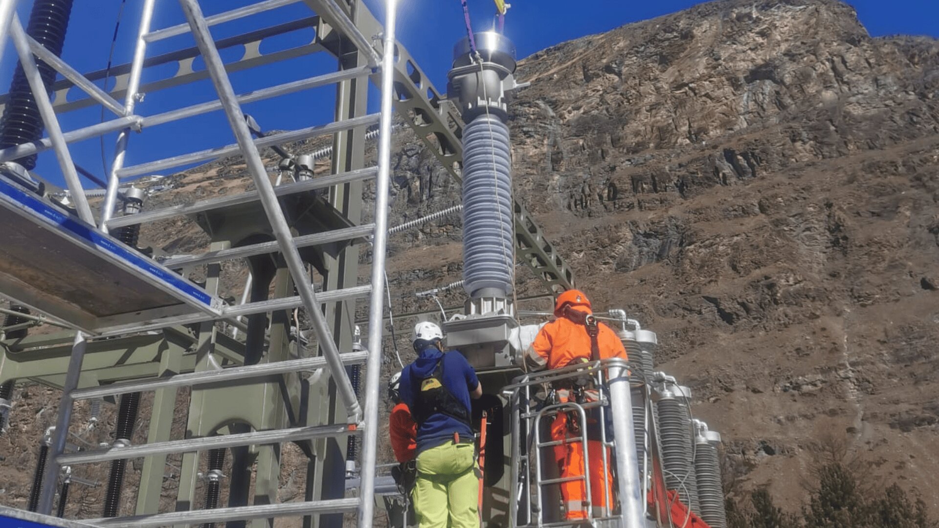 Zwei Arbeiter in Schutzkleidung stehen auf einer Hebebühne und führen Wartungsarbeiten an elektrischen Hochspannungsgeräten im Freien durch. Im Hintergrund ist ein felsiger Berghang bei strahlend blauem Himmel zu sehen.