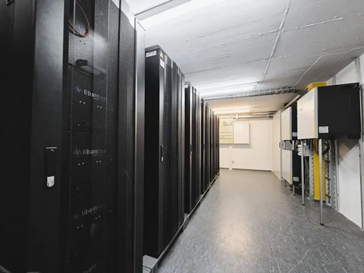 Eine Reihe schwarzer Serverschränke in einem hell erleuchteten, modernen Rechenzentrumsraum mit weißen Wänden und elektronischen Schalttafeln an der rechten Seite.