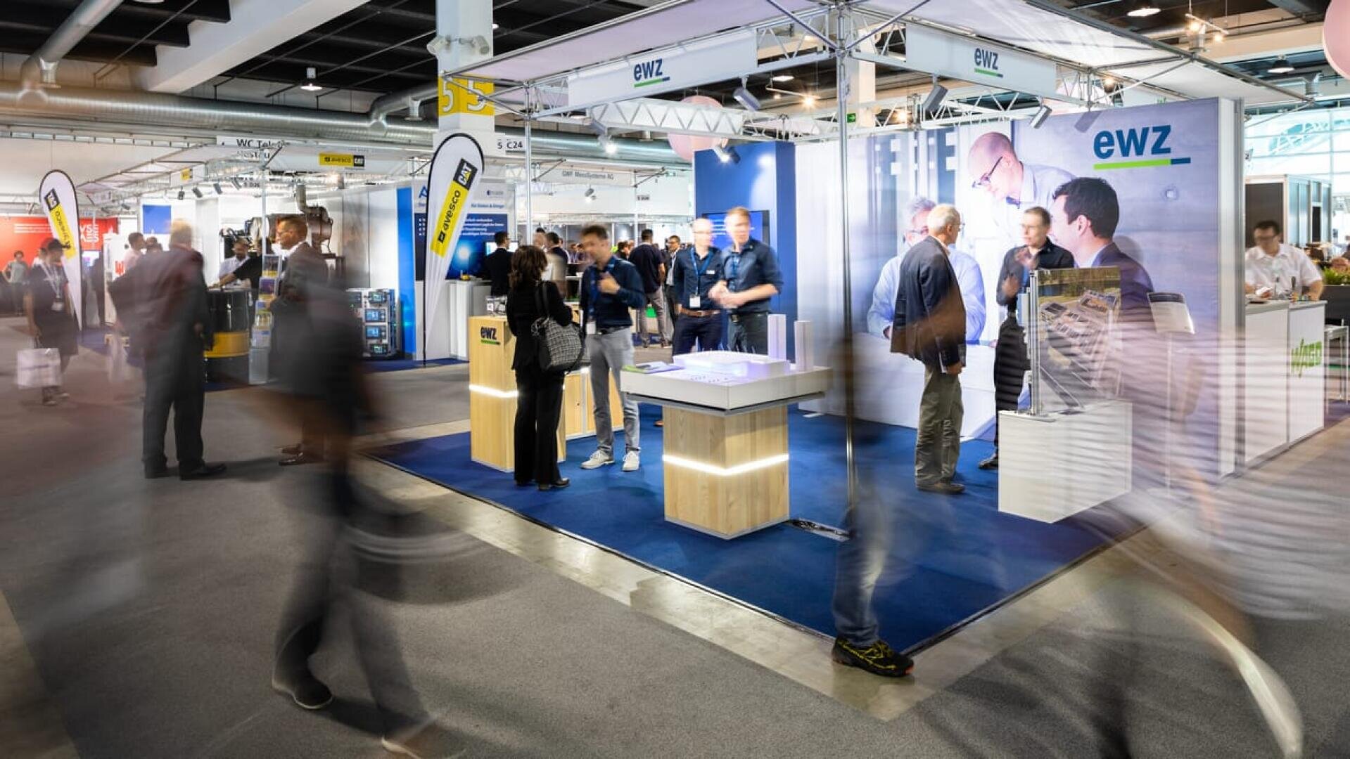 Eine belebte Messe mit Menschen, die an einem hell erleuchteten Stand mit der Aufschrift ewz vorbeilaufen. Mehrere Personen unterhalten sich im Inneren des Standes, und im gesamten Veranstaltungsbereich sind Informationsdisplays zu sehen.