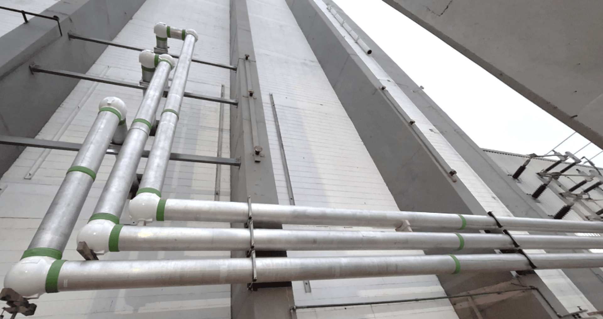 Drei parallele silberne Metallrohre mit grünen Markierungen verlaufen senkrecht und waagerecht entlang der Außenwand eines hohen Gebäudes und werden von Metallbügeln gehalten. Das Foto ist aus einem niedrigen Winkel aufgenommen, mit Blick nach oben.
