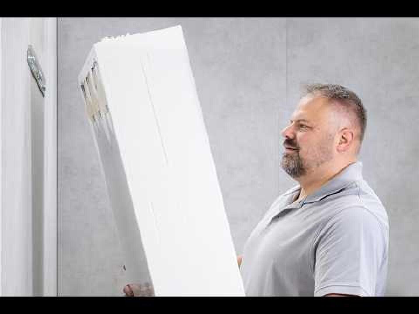 Ein Mann mit grauem Bart und hellgrauem Polohemd hebt einen großen weißen rechteckigen Gegenstand, möglicherweise ein Möbelstück, in einem Raum mit hellgrauen Wänden hoch und untersucht ihn.