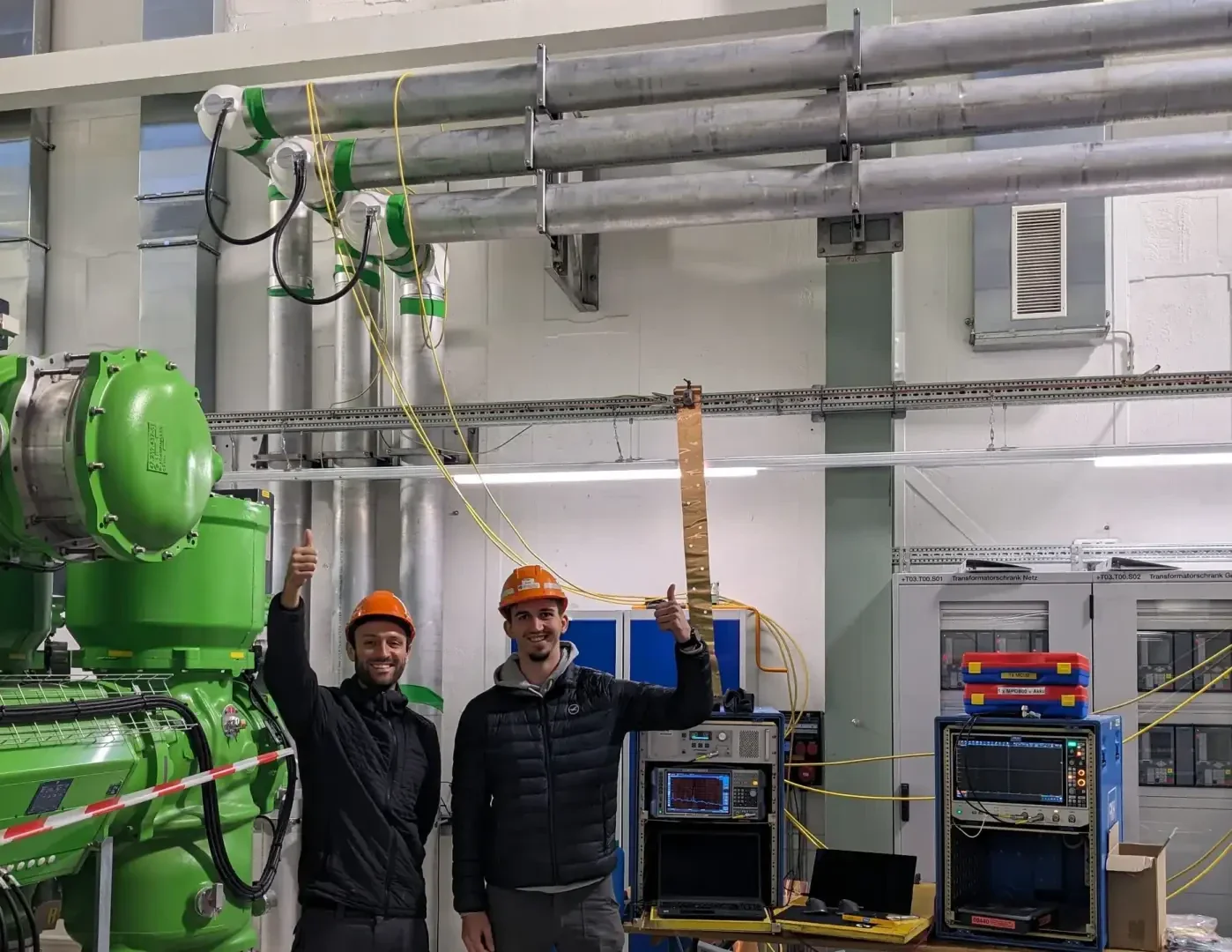 Zwei Personen mit orangefarbenen Schutzhelmen und schwarzen Jacken stehen in einem Industrielabor, lächeln und zeigen die Daumen nach oben. Einer hält ein langes Kupferband, im Hintergrund sind Geräte, Kabel und Maschinen zu sehen.