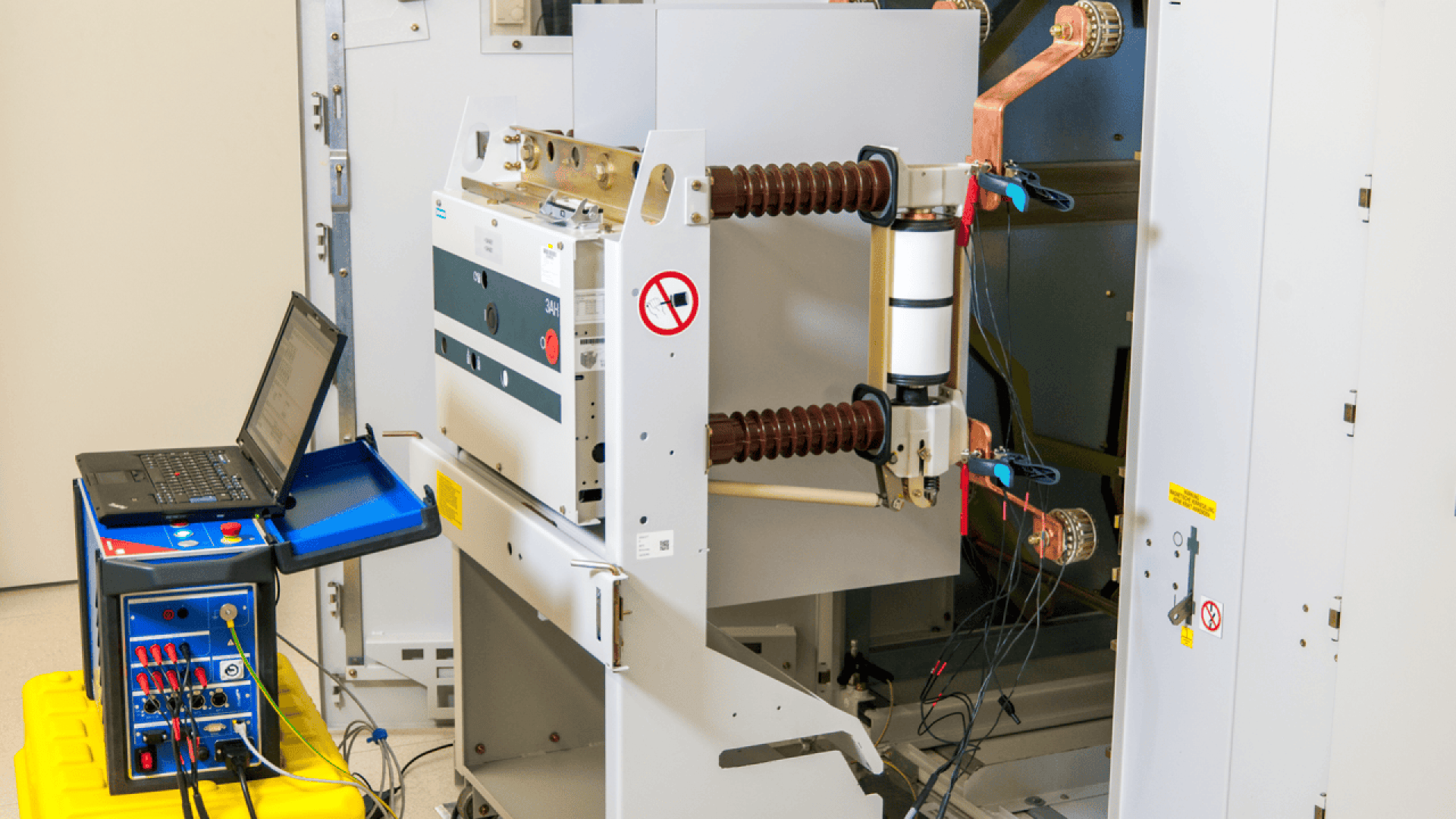 A high-voltage electrical circuit breaker is being tested with diagnostic equipment; cables and probes are connected, and a laptop is recording data. Safety signs are visible on the breaker.