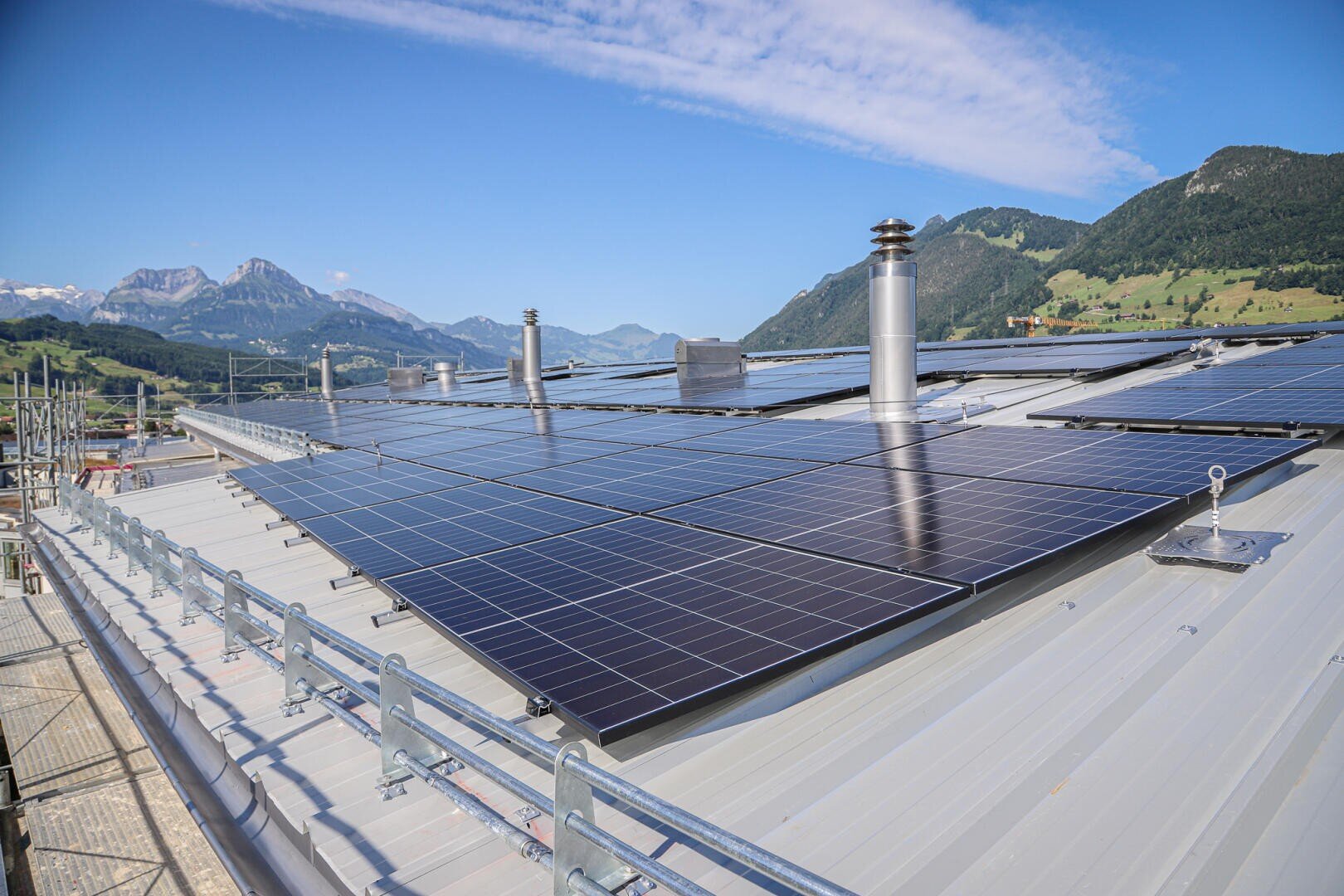 Reihen von Solarmodulen, die auf einem Metalldach installiert sind, mit Schornsteinen und Bergen im Hintergrund unter einem strahlend blauen Himmel mit vereinzelten Wolken.