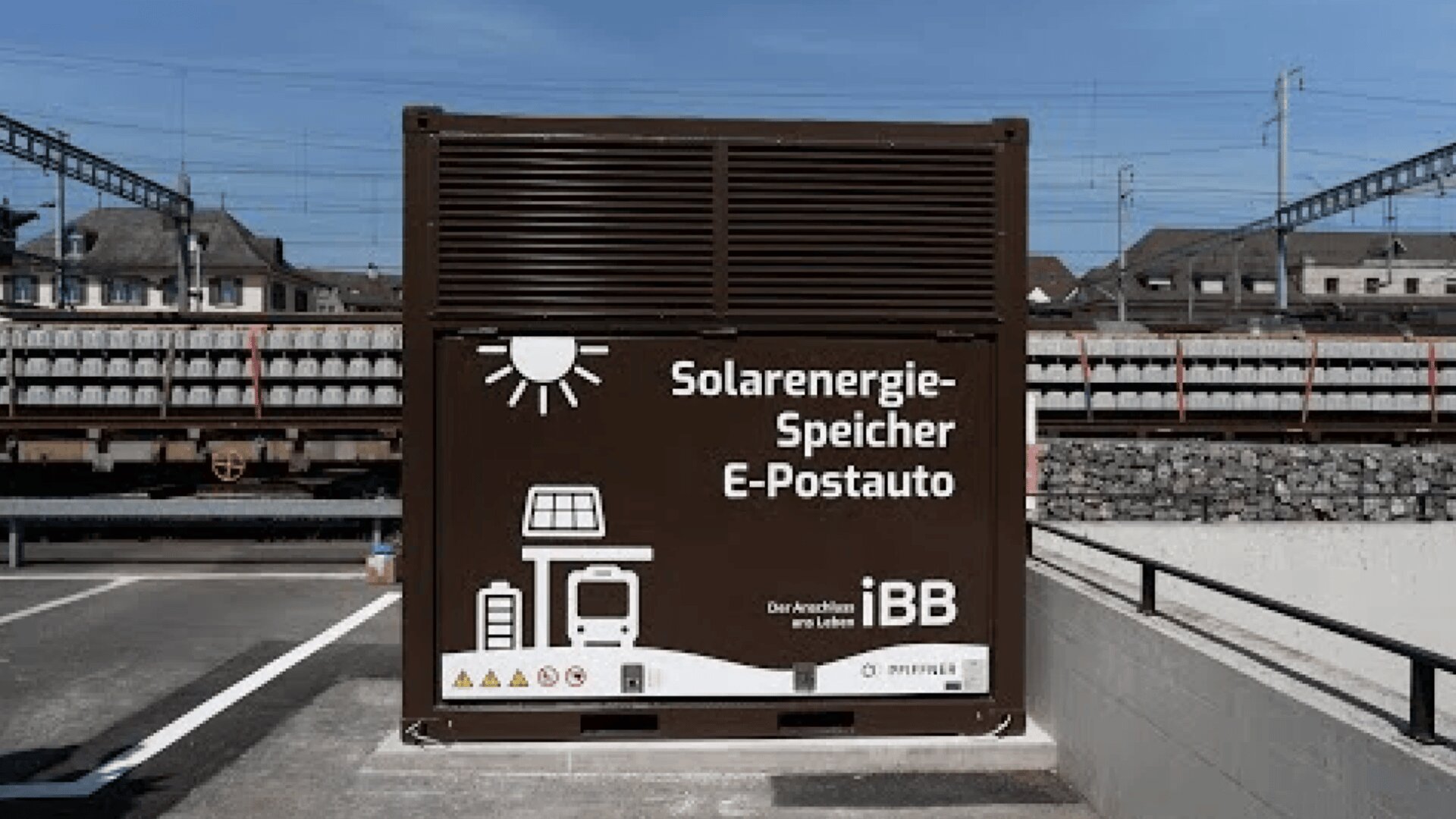 Large brown container labeled “Solarenergie-Speicher E-Postauto” with white icons depicting solar panels, a bus, and charging. It is situated outdoors near train tracks and buildings under a clear sky.