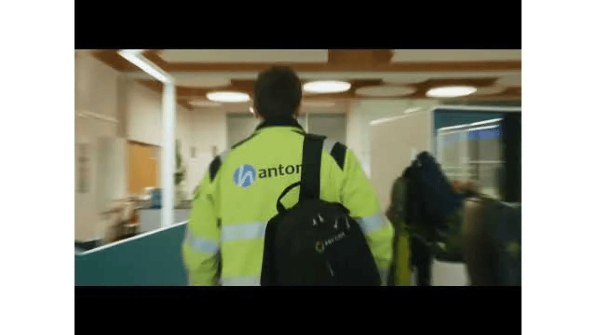 A person wearing a high-visibility jacket with antor on the back walks through an office carrying a black backpack. The scene shows an indoor workspace with desks and partition walls.
