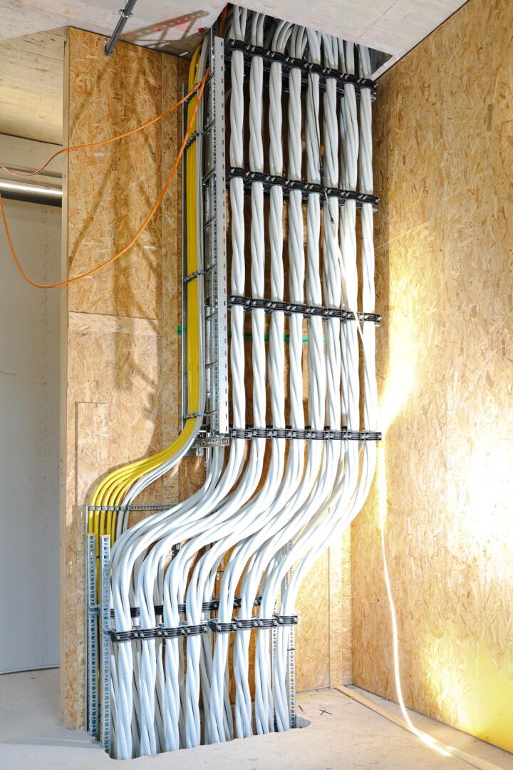 Ein ordentlich geordnetes Bündel weißer und gelber Elektrokabel verläuft vertikal an einer Wand hinauf und ist mit Metallklammern in einem modernen Gebäude mit freiliegenden Wänden aus Litzendielen befestigt.