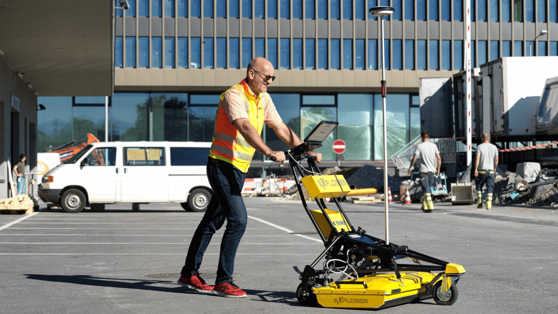 Ein Mann in einer Sicherheitsweste benutzt ein gelbes Bodenradargerät auf einem Parkplatz in der Nähe einer Baustelle, mit Arbeitern, Fahrzeugen und einem modernen Gebäude im Hintergrund.