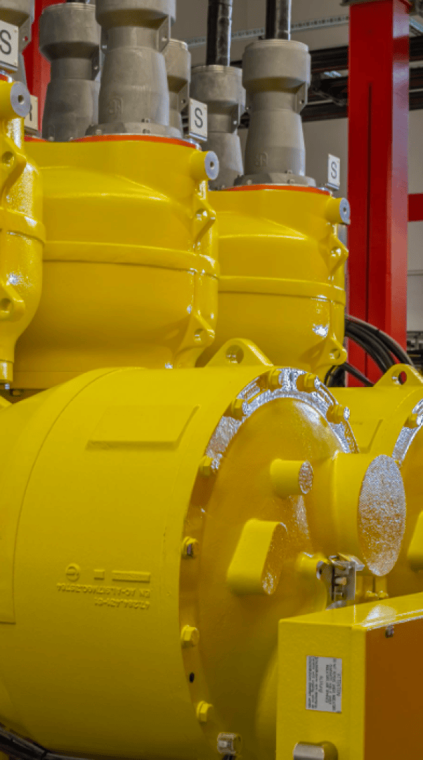 Nahaufnahme einer leuchtend gelben Industriemaschine mit Bolzen und zylindrischen Bauteilen vor einem unscharfen Hintergrund mit roten vertikalen Balken und grauen Beschlägen.
