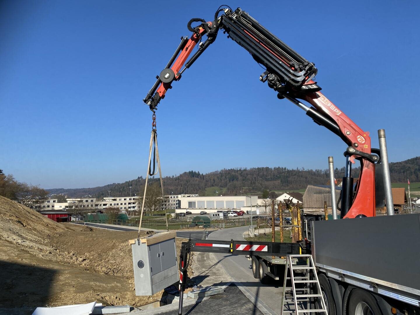 Ein auf einem Lastwagen montierter Kran hebt an einem klaren Tag eine große Metallbox auf einer Baustelle an. Im Hintergrund sind Gebäude, ein Hügel und einige Bäume unter einem blauen Himmel zu sehen. Eine Metallleiter lehnt an dem Lastwagen.