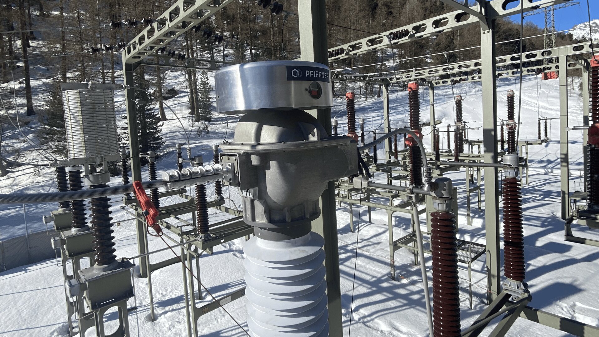 Elektrisches Umspannwerk im Freien in einer verschneiten Landschaft mit Metallstrukturen, Isolatoren und verschiedenen elektrischen Bauteilen mit Bergen und Bäumen im Hintergrund unter einem klaren Himmel.