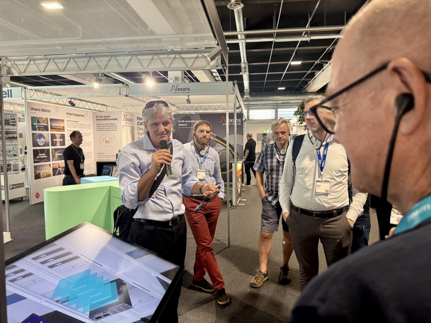 Ein Mann, der ein Mikrofon in der Hand hält, präsentiert einer Gruppe von Besuchern auf einer Technologie- oder Fachmesse Informationen auf einem großen Touchscreen, während im Hintergrund Messestände und Ausstellungsstücke zu sehen sind.