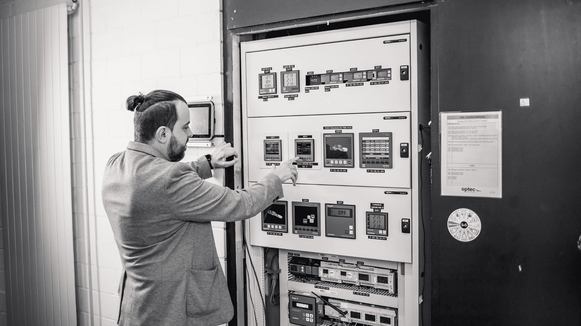 A man in a suit operates a large control panel with multiple switches, buttons, and screens in an industrial or technical setting.