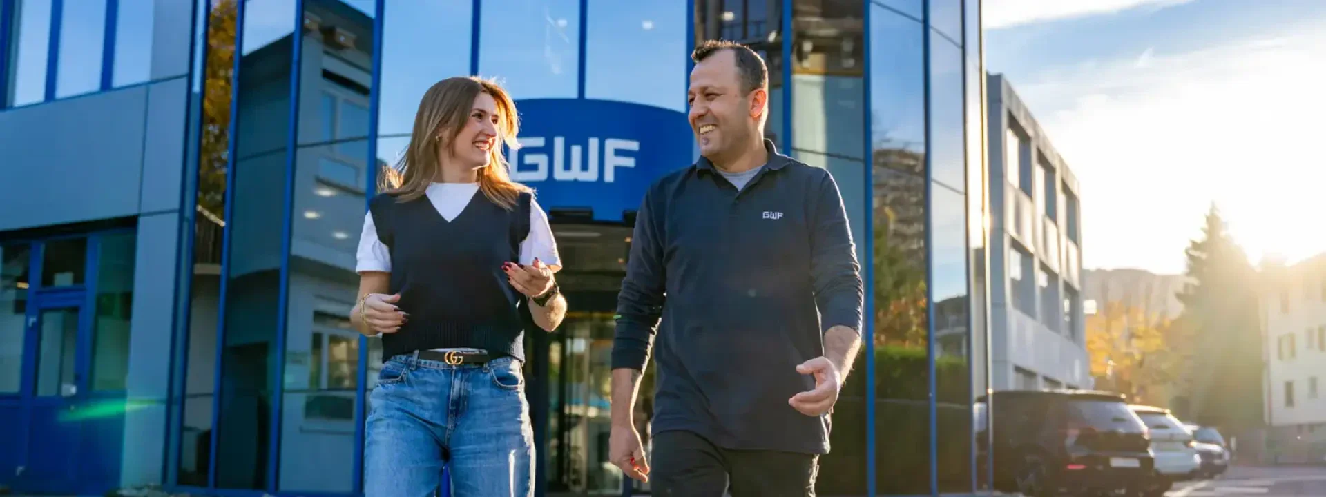 A woman and a man are smiling and chatting as they walk in front of a modern glass office building with a sign above the entrance that reads GWF. The sun is shining and the atmosphere is friendly and informal.