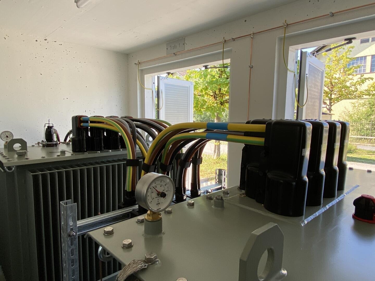 Eine Nahaufnahme von elektrischen Transformatoren und bunten Kabeln in einem Raum mit zwei offenen Fenstern, die Grün und Sonnenlicht nach draußen lassen. An den Geräten sind ein Manometer und Kühlerlamellen zu sehen.