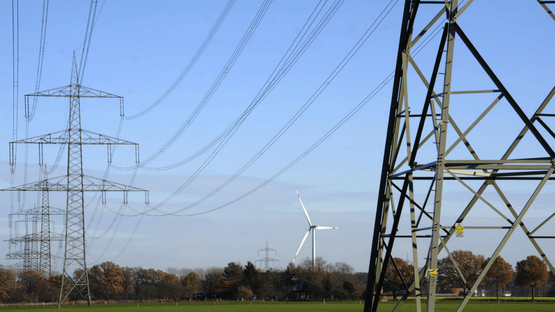 Hochspannungsleitungen und Metallmasten erstrecken sich über eine grüne Wiese mit Bäumen, und im Hintergrund ist eine Windturbine unter blauem Himmel zu sehen.