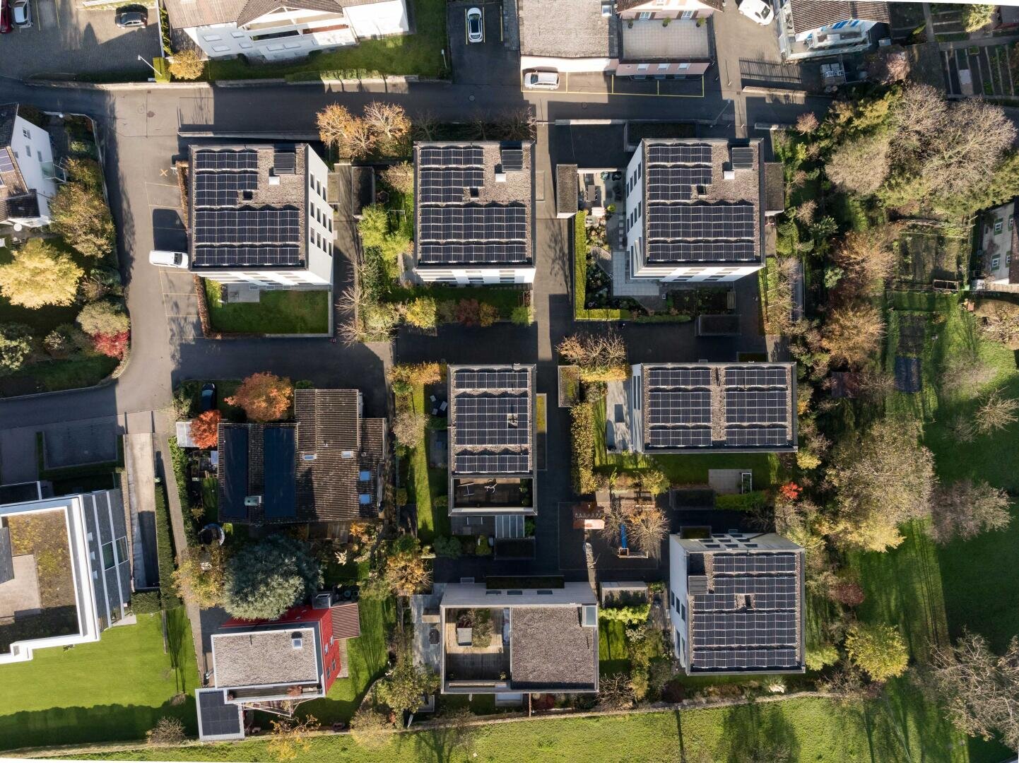 Luftaufnahme eines Wohnviertels mit mehreren modernen Gebäuden, jedes mit Solarzellen auf den Dächern, umgeben von grünen Rasenflächen, Bäumen und sich kreuzenden Straßen.