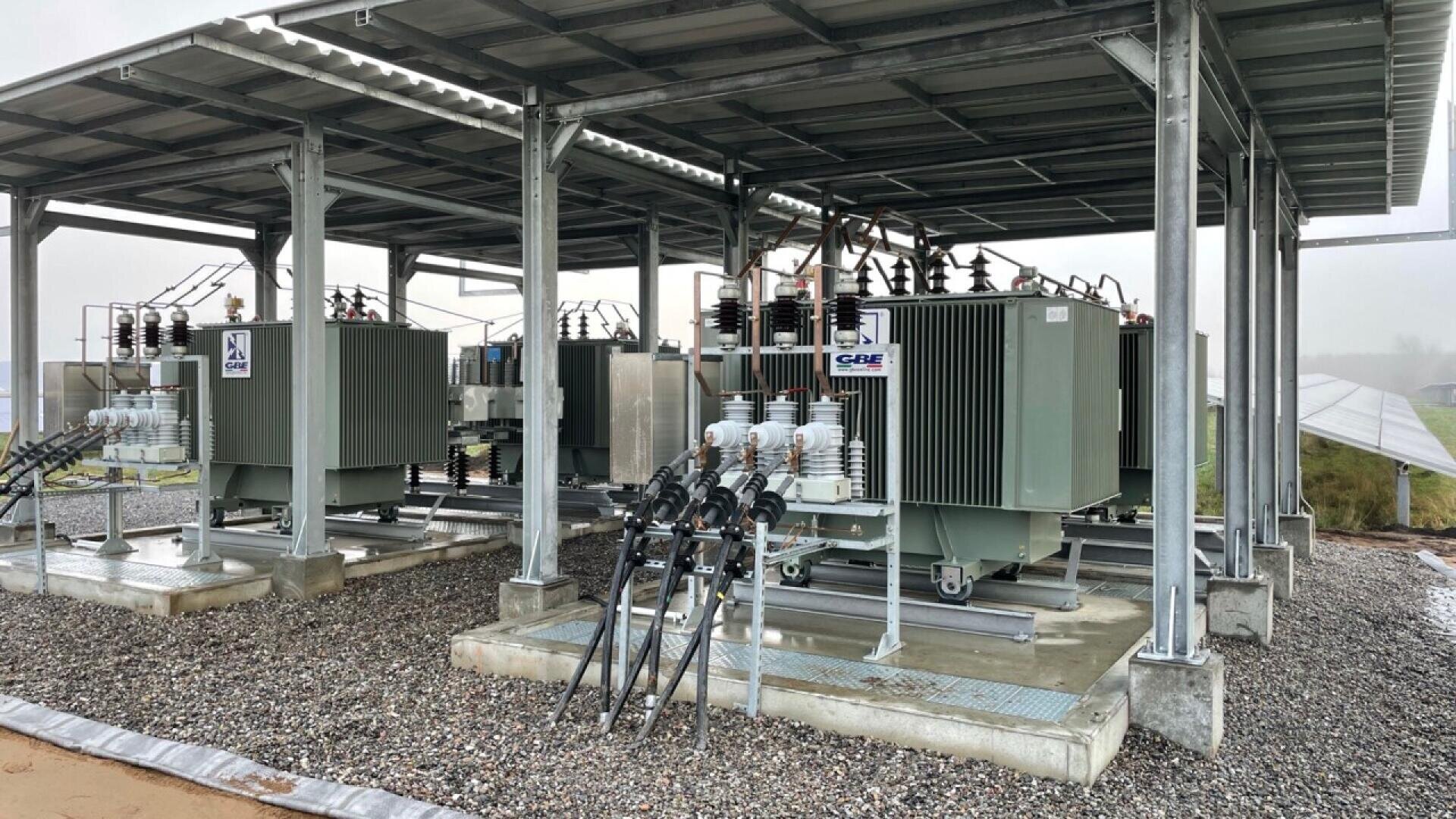 Zwei große Stromtransformatoren sind unter einem Metalldach auf einer Kiesfläche installiert, wobei die Kabel zu jedem Gerät und die Schalttafeln sichtbar sind; der Hintergrund ist ein grüner Außenbereich.