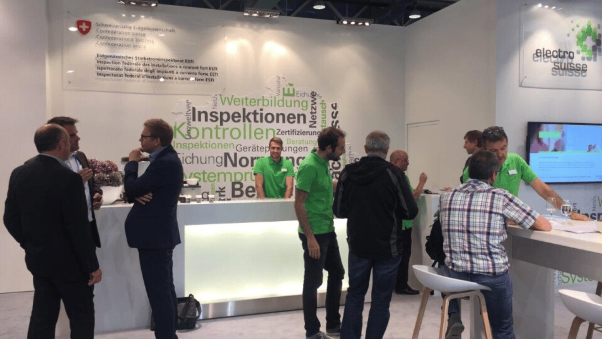 Several people talk and network at a trade show booth for Electrosuisse, with a large word cloud and informational text on the wall behind them. Some staff members wear matching green shirts. A digital screen displays information.