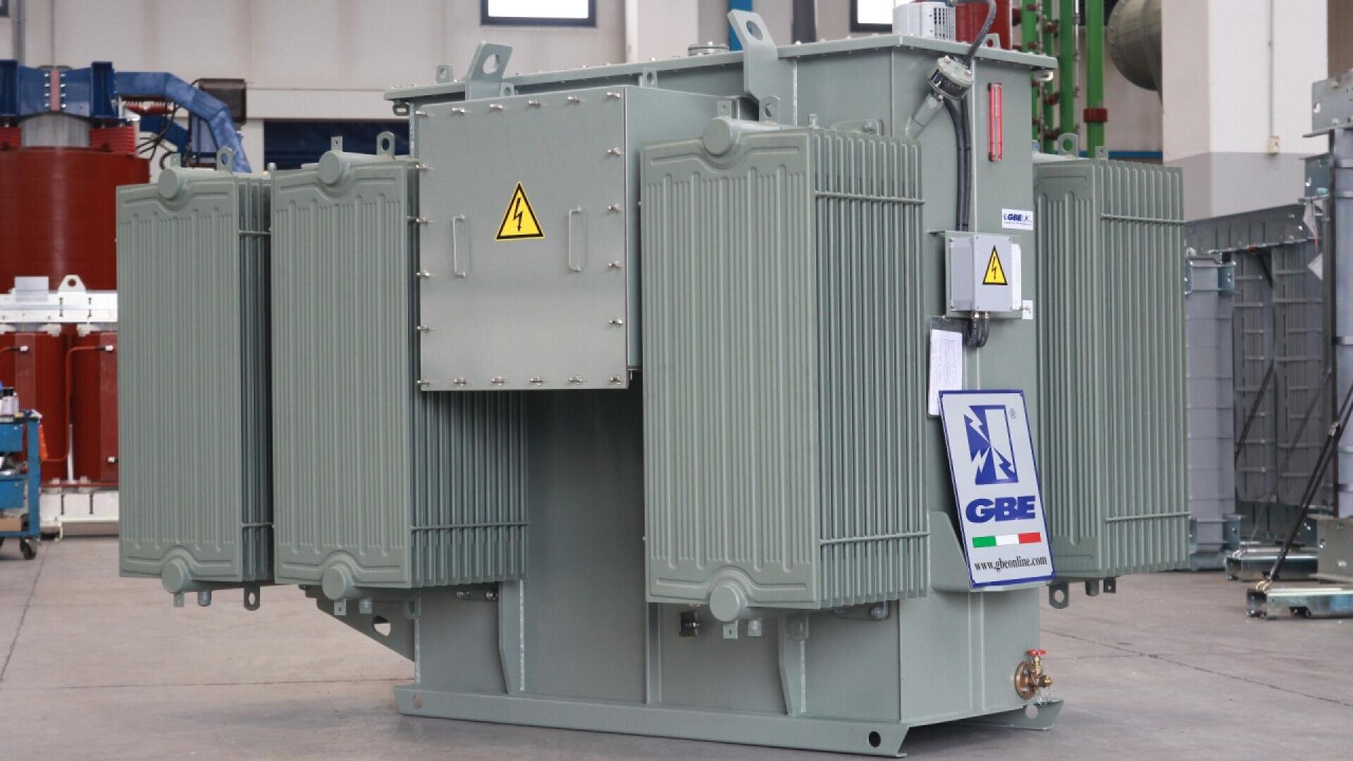 Ein großer elektrischer Industrietransformator mit Kühlrippen aus Metall ist in einem Innenraum ausgestellt. Es sind Warnschilder und ein GBE-Firmenlogo auf der Vorderseite zu sehen. Im Hintergrund sind Industrieanlagen zu sehen.