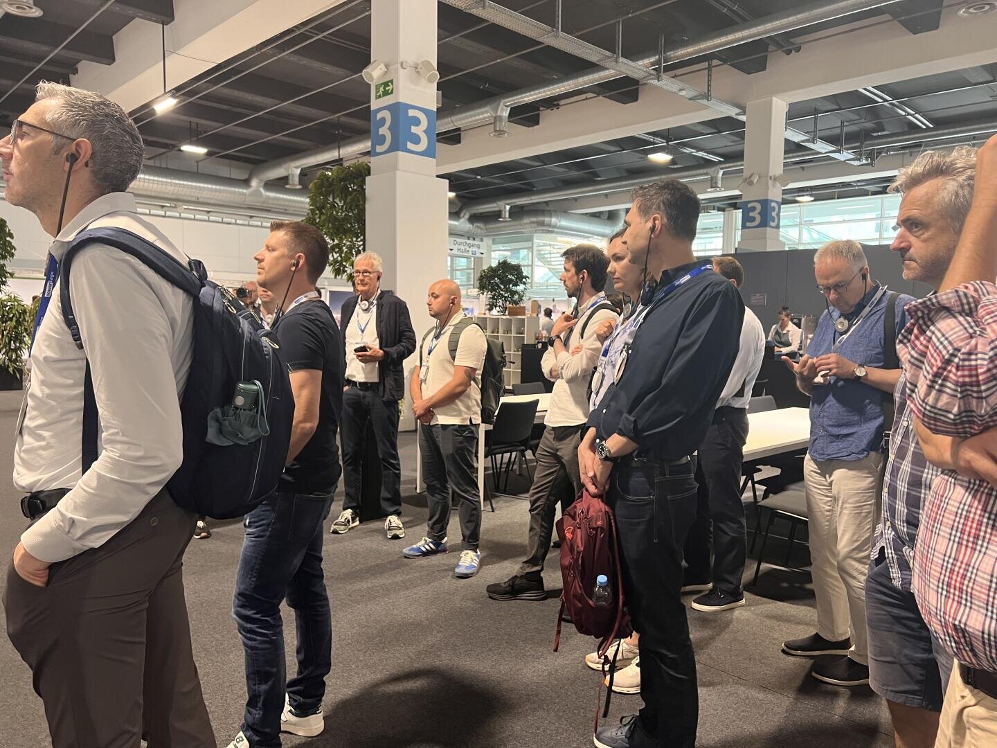 Eine Gruppe von Menschen, überwiegend Männer, steht aufmerksam in einem Raum bei einer Veranstaltung oder Konferenz. Einige tragen Namensschilder und haben Rucksäcke oder Taschen dabei. Im Hintergrund sind nummerierte Schilder und Tische zu sehen.