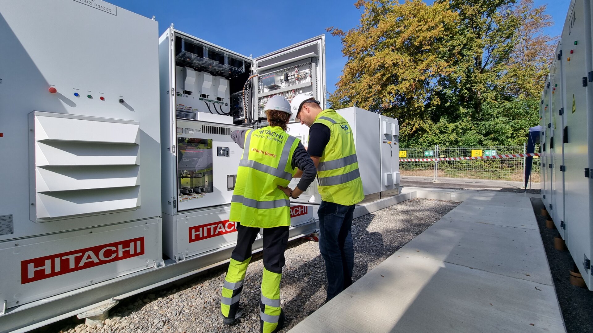 Zwei Arbeiter mit Sicherheitswesten und Helmen inspizieren an einem sonnigen Tag im Freien eine offene elektrische Schalttafel, während im Hintergrund HITACHI-Geräte und Bäume zu sehen sind.