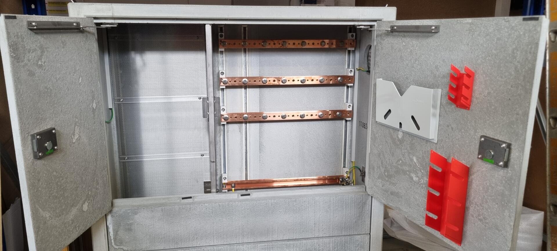 Ein offener Schaltschrank mit drei horizontalen Kupferschienen im Inneren, grauen Metalltüren und roten Kunststoffteilen an der rechten Innentür.